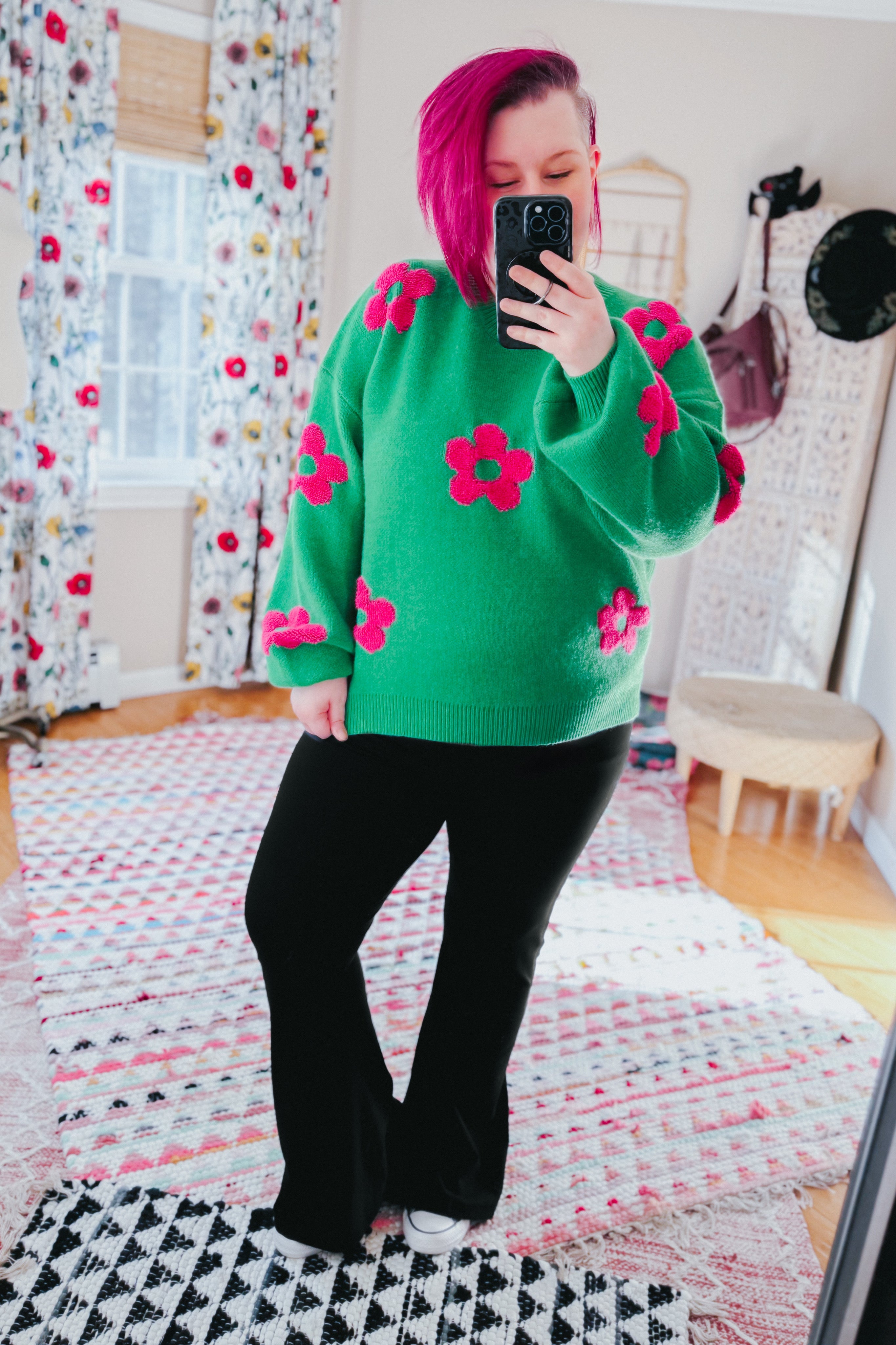 Woman wearing a green sweater embellished with tufted pink flowers and black yoga pants in an indoor setting.