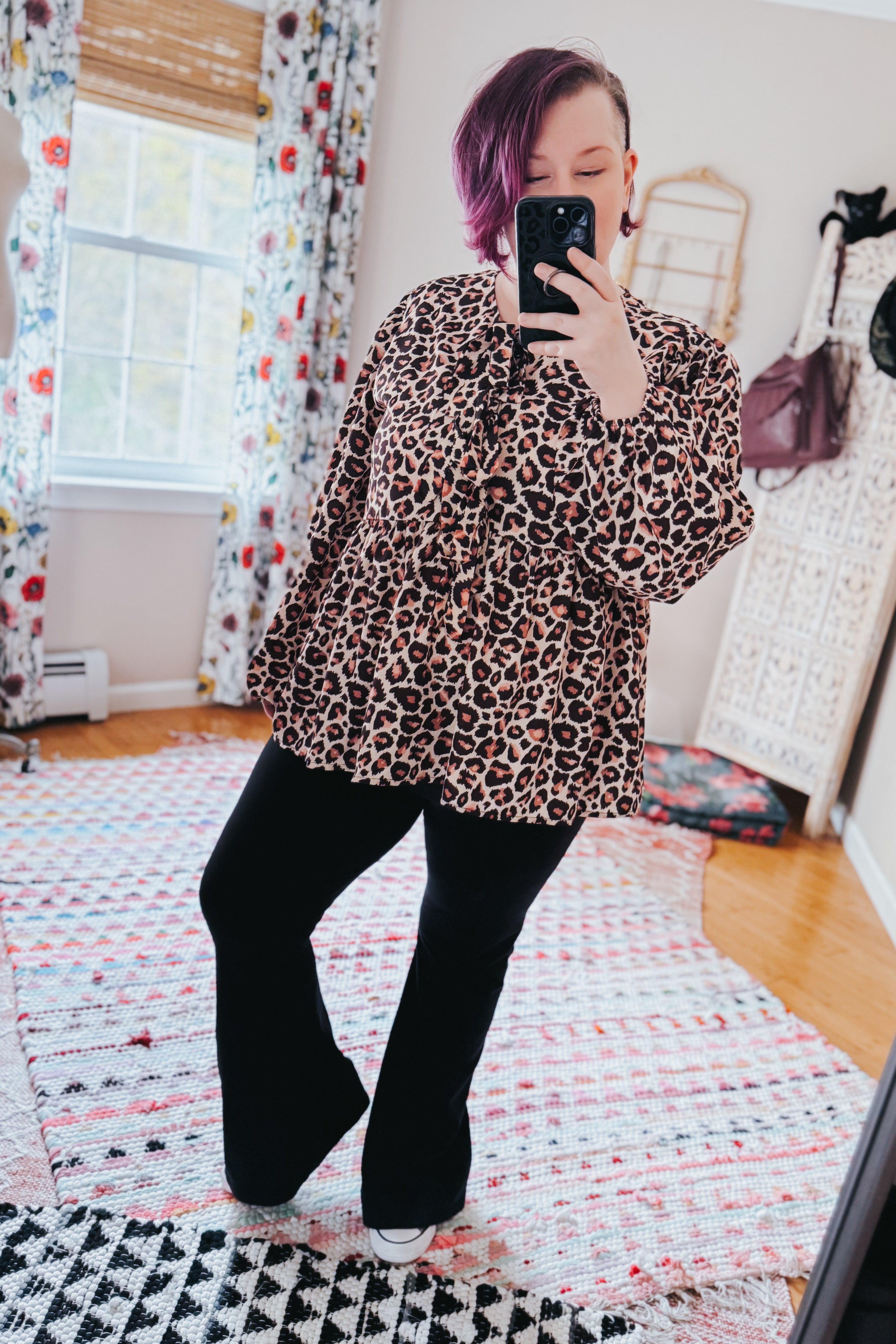 Woman taking mirror selfie wearing brown leopard print babydoll blouse with long sleeves and black flared pants in bright room