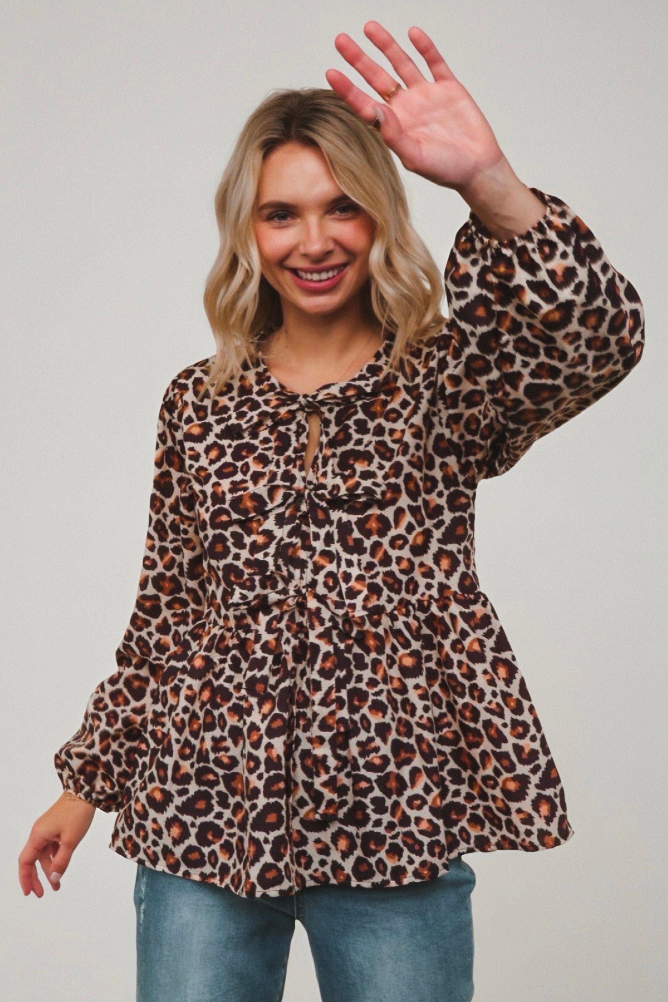Smiling woman wearing brown leopard print babydoll blouse with long balloon sleeves and ruffle details paired with jeans waving hand