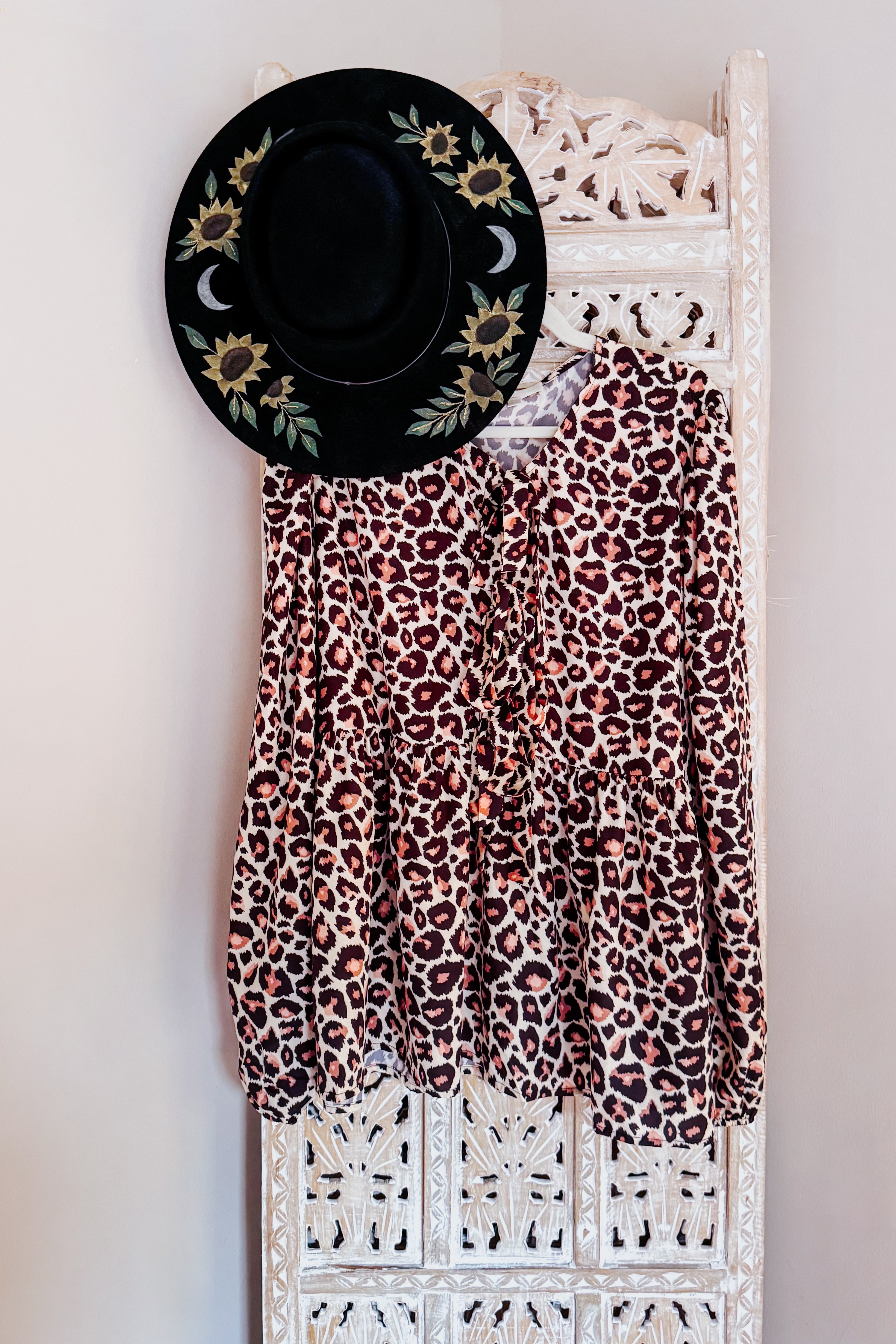 Brown leopard print babydoll blouse with long sleeves displayed on carved white wooden screen alongside black floral hat