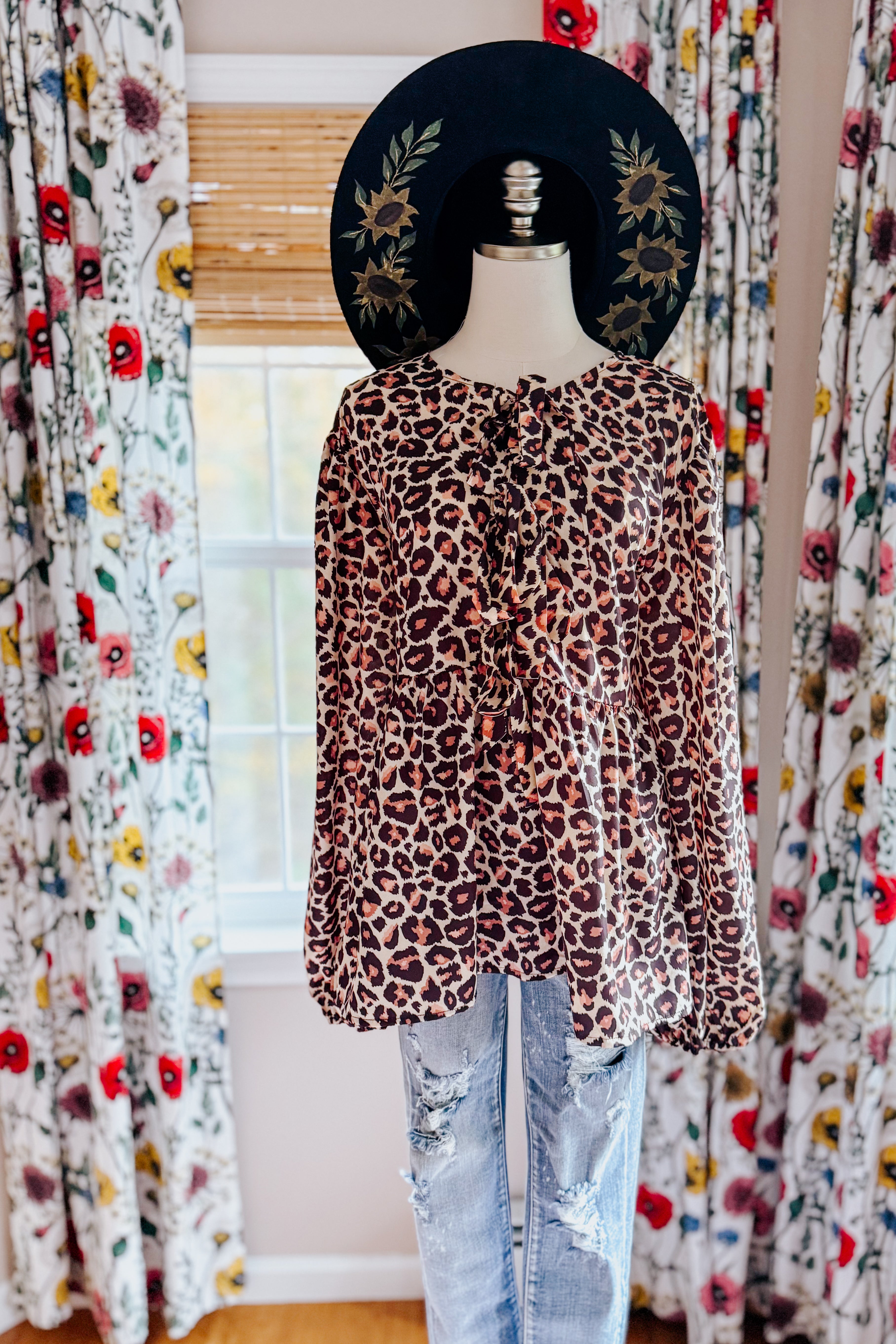 Brown leopard print babydoll blouse with long sleeves styled on mannequin with distressed jeans and black floral hat
