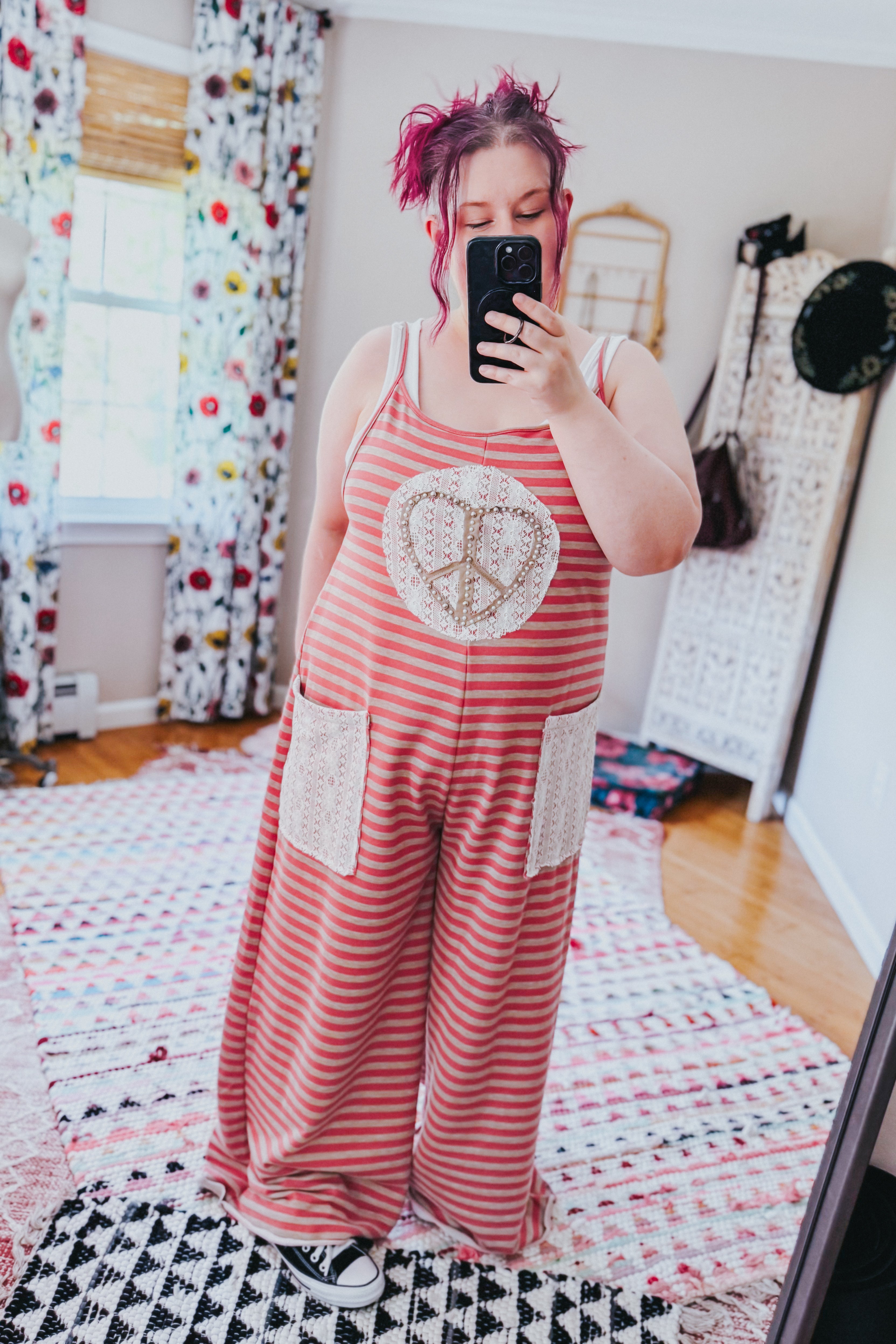 Woman taking mirror selfie wearing loose red striped jumpsuit with lace patch pockets and peace sign detail on front