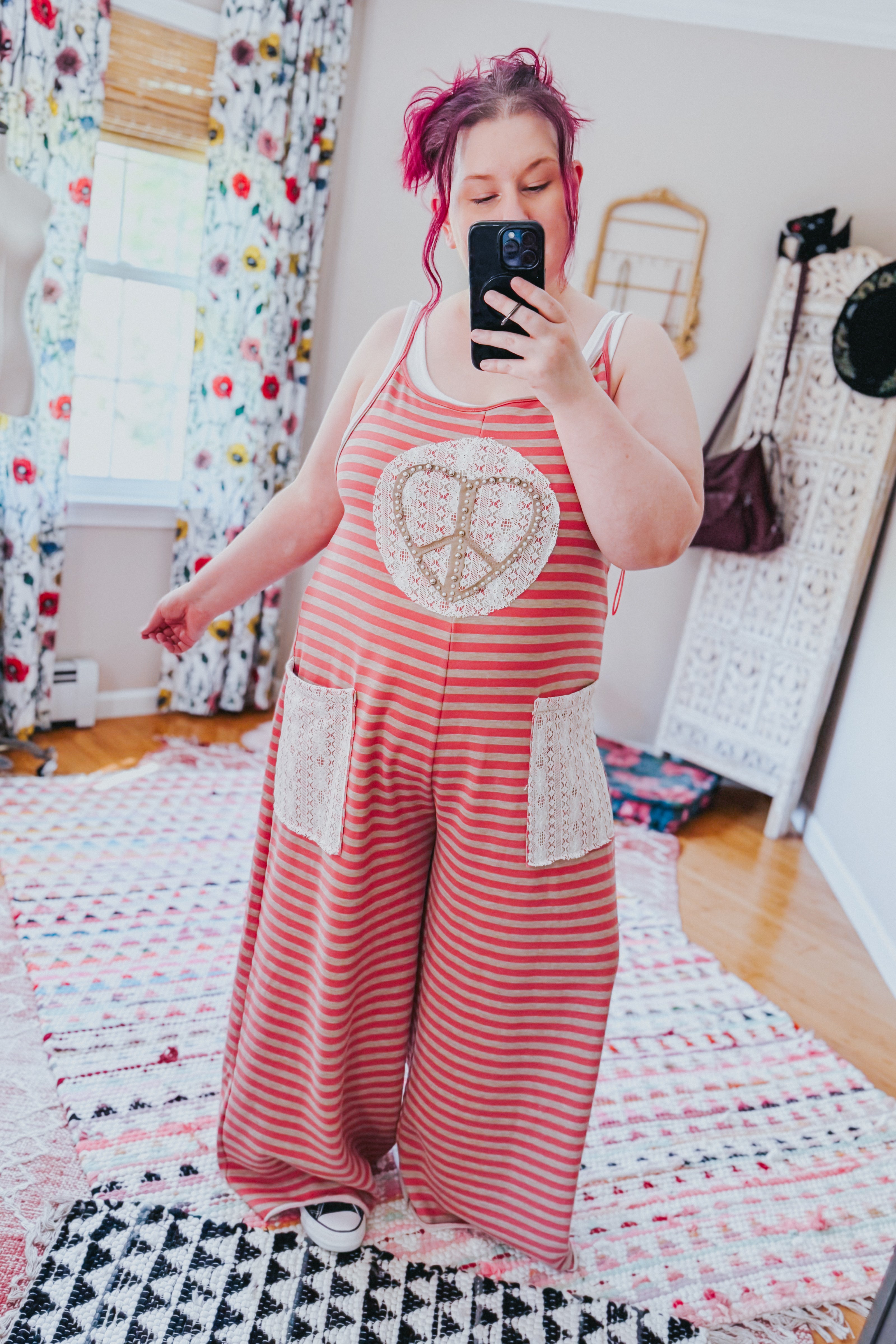 Woman wearing loose red striped jumpsuit with lace pockets and large peace sign applique taking selfie in colorful room