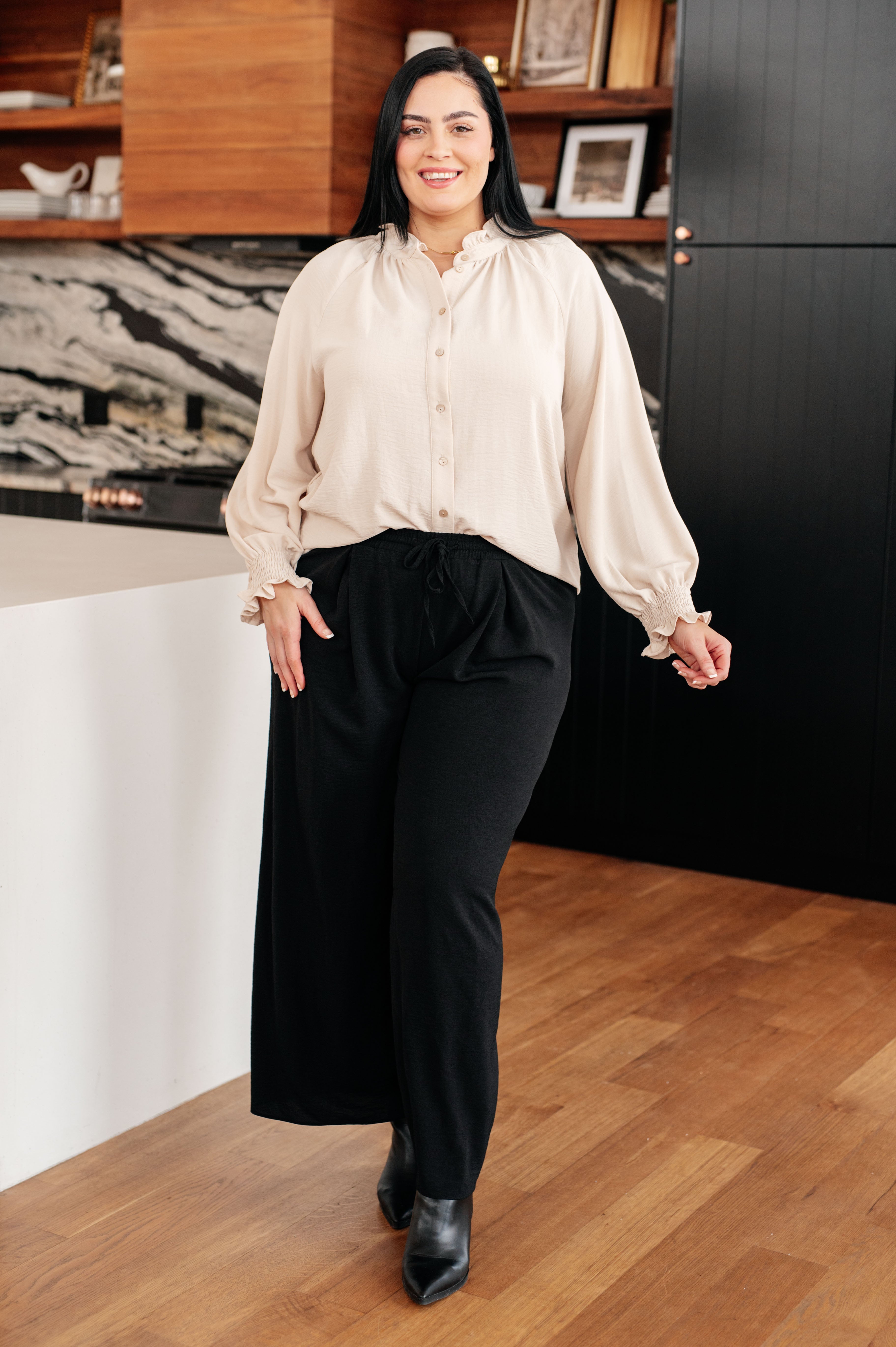 Woman wearing cream button-up blouse with ruffled neckline and smocked ruffle cuffs paired with black wide-leg pants