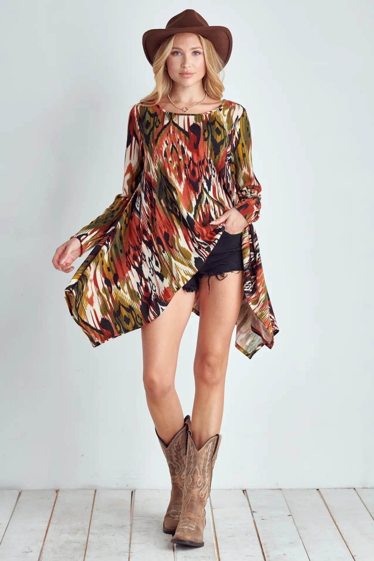 Woman wearing flowing multicolor abstract patterned shark bite hem top with black shorts, cowboy boots, and brown hat against plain background
