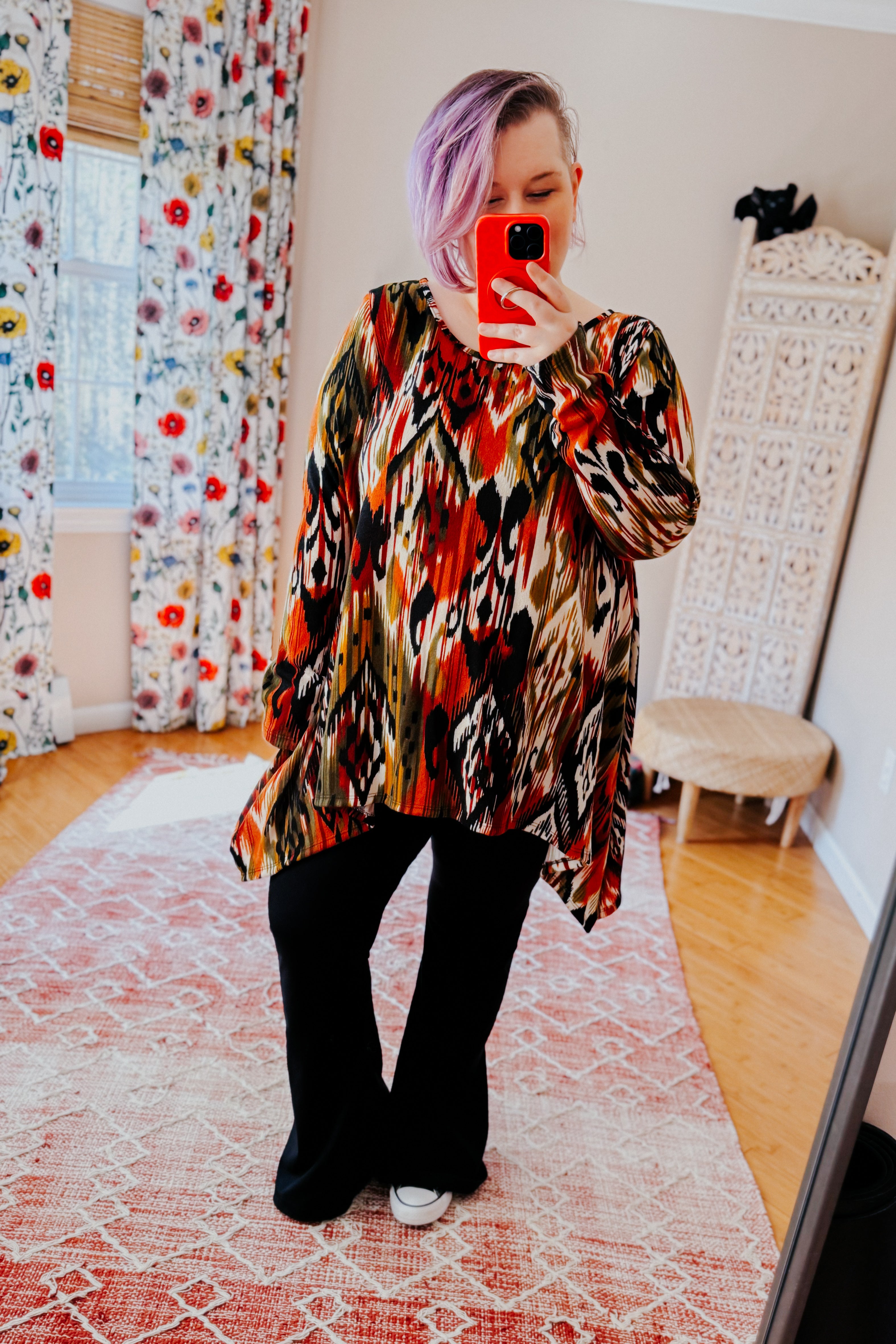 Woman with light purple hair wearing colorful red and green patterned long sleeve asymmetrical top and black pants indoors taking selfie