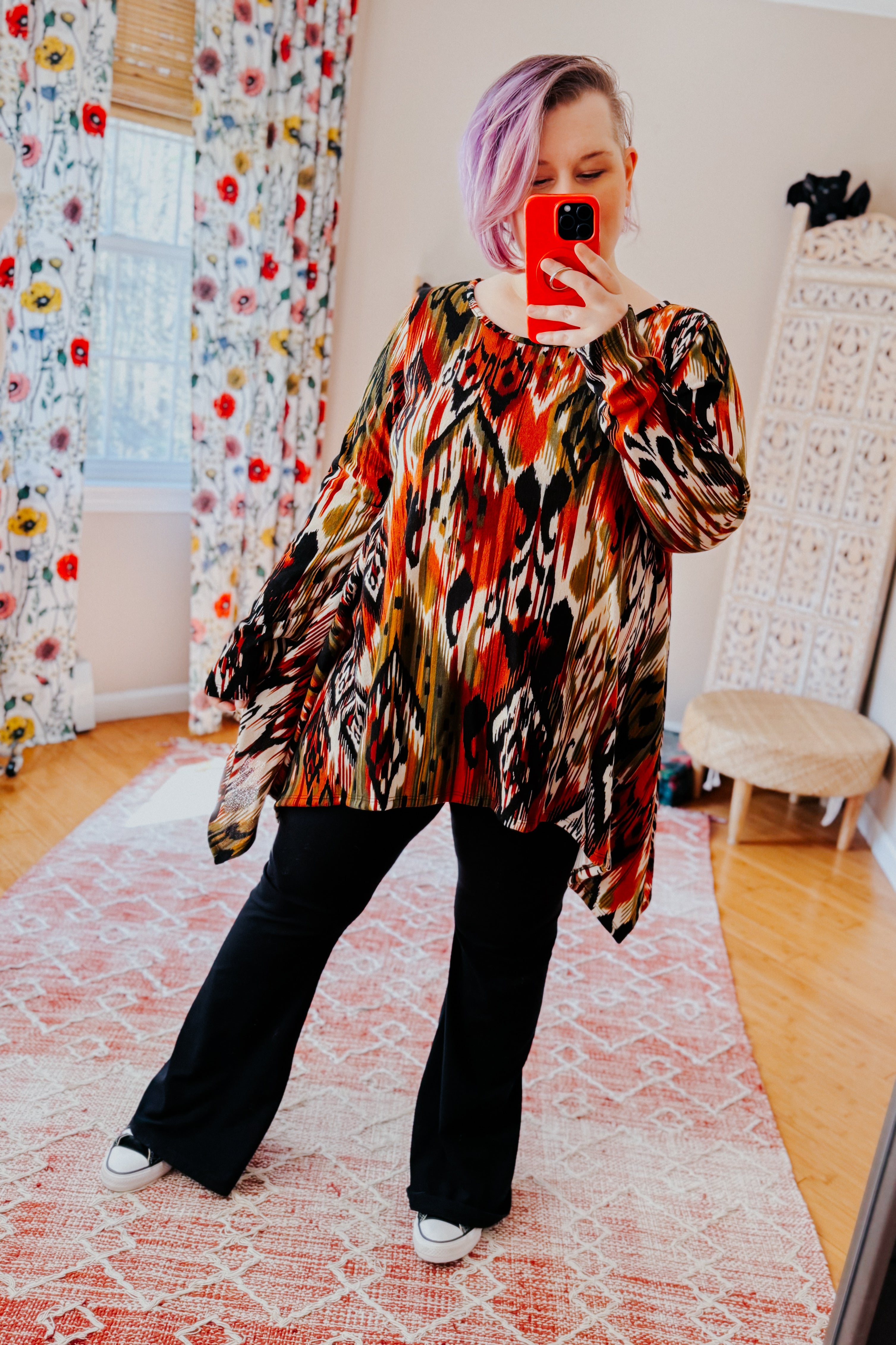 Woman with light purple hair wearing red and black abstract patterned flowing top with black pants in bright room taking selfie