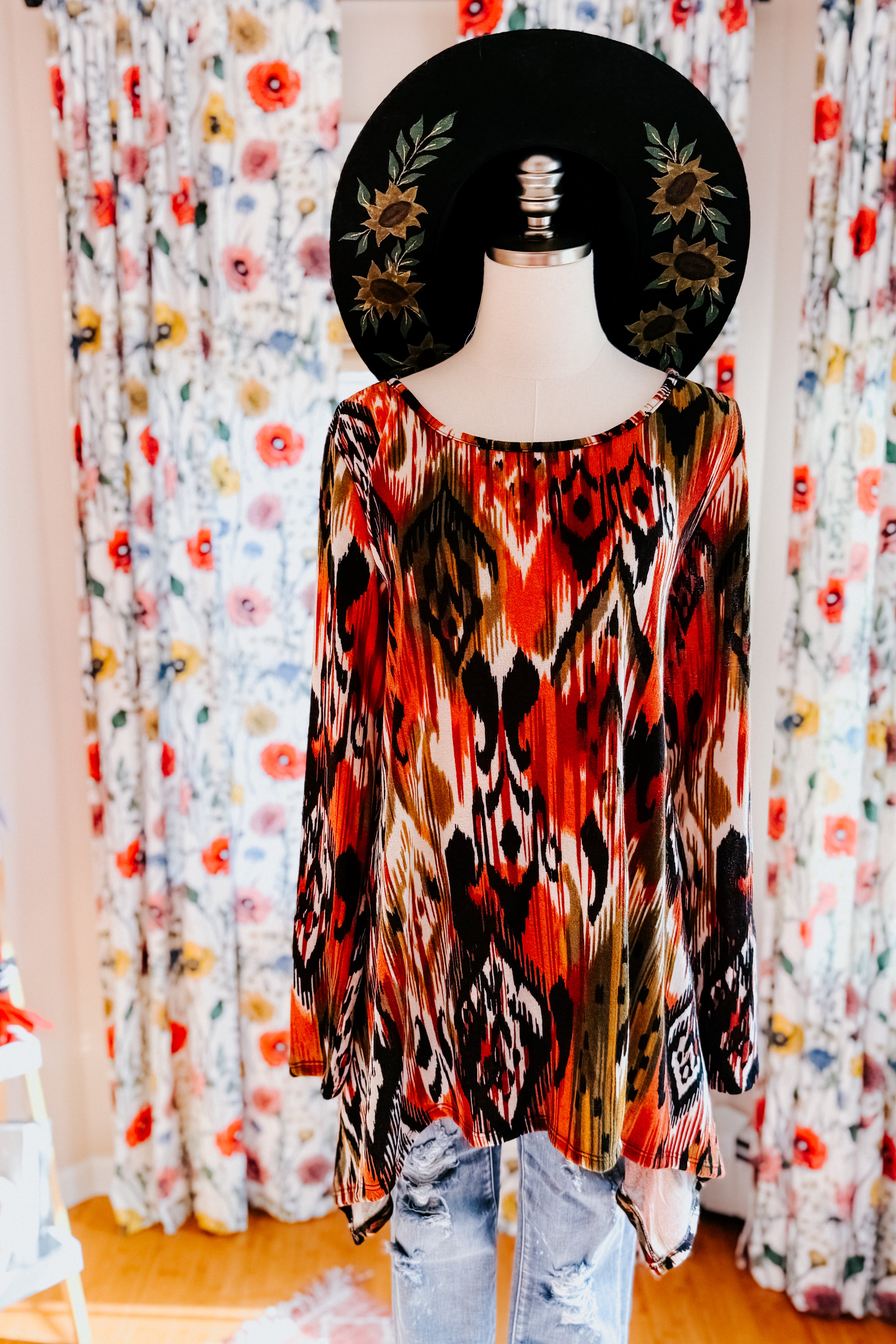 Mannequin wearing red, orange, and black patterned flowing long sleeve top paired with distressed light blue jeans and black wide-brimmed hat