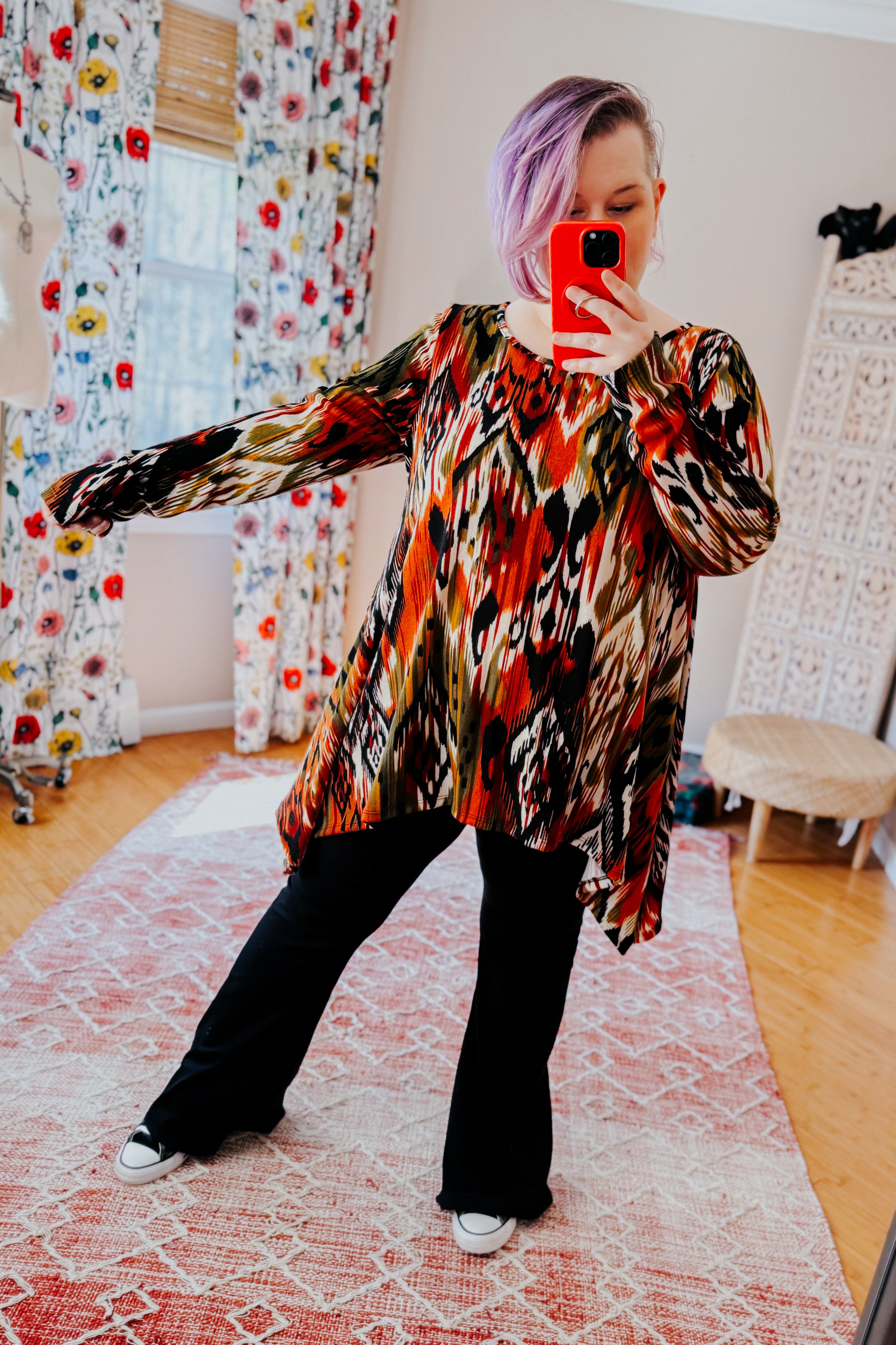 Woman with light purple hair wearing red, green, and black patterned long sleeve asymmetrical top with black pants indoors taking selfie