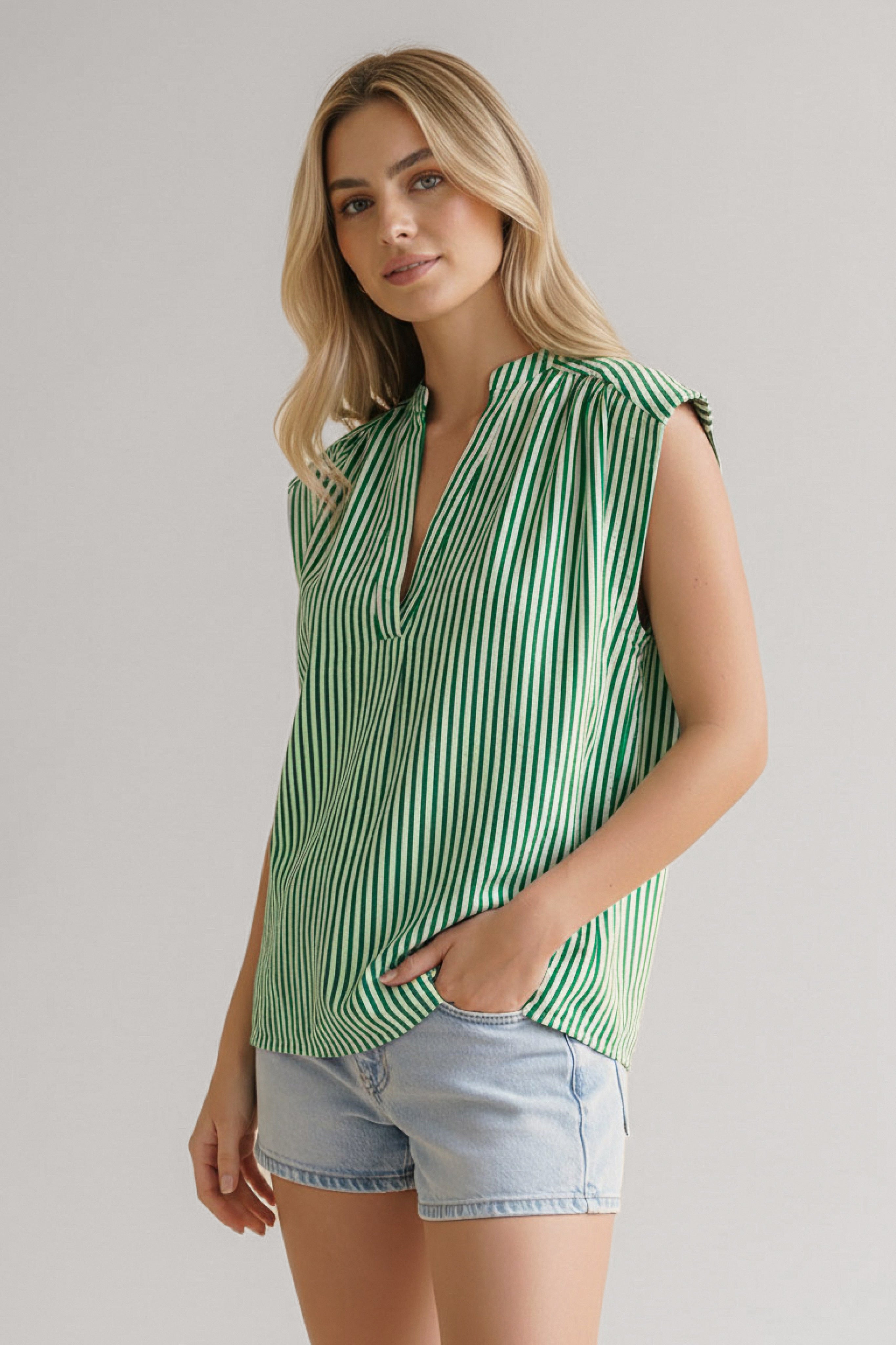 Woman wearing green striped sleeveless blouse with V-neck paired with light denim shorts standing against neutral background