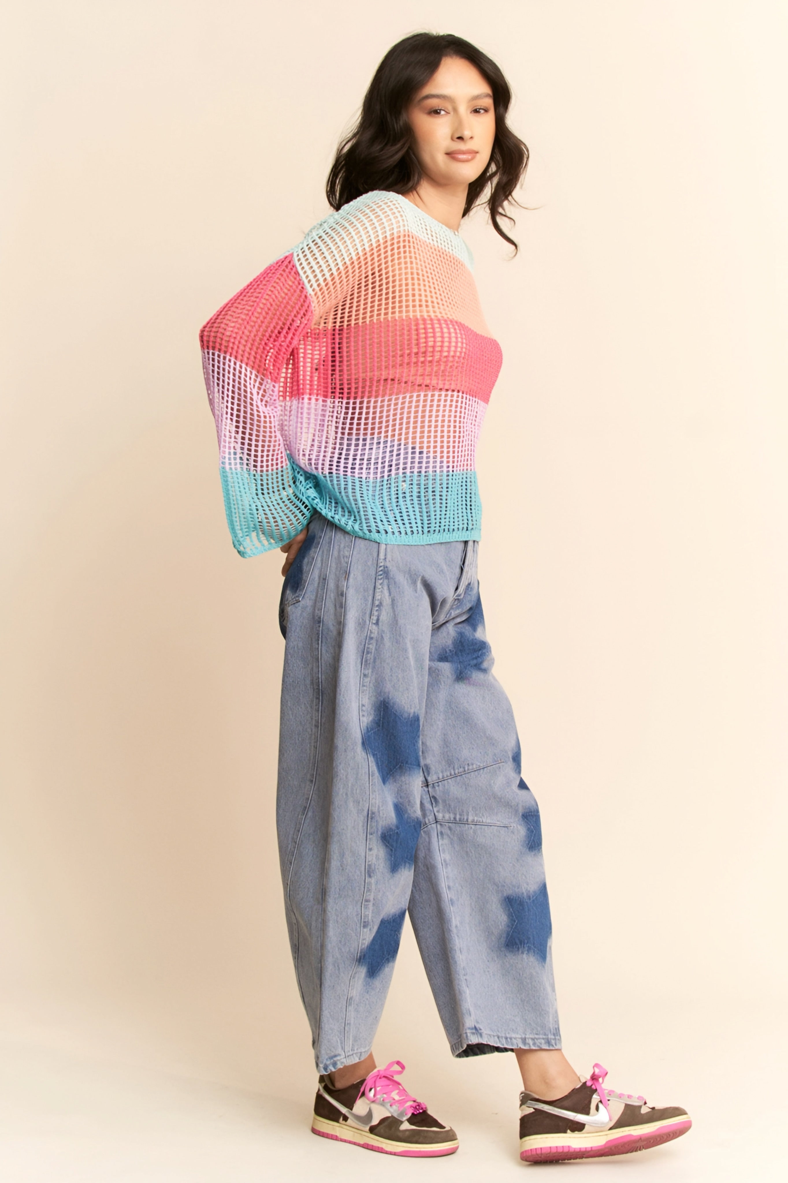 Woman wearing multicolor striped crochet mesh pullover with bell sleeves paired with wide-leg tie-dye jeans and sneakers