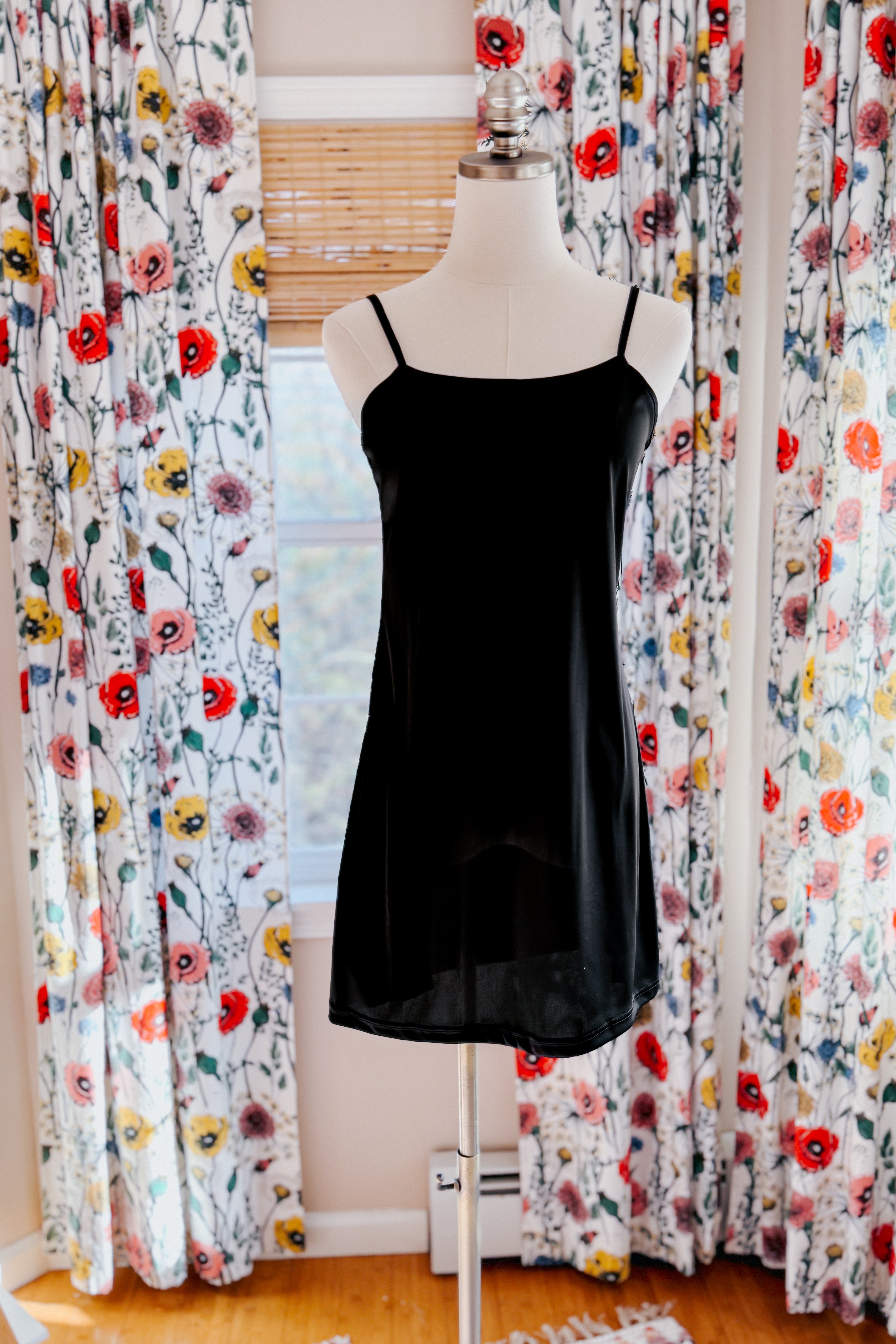 Black slip dress with thin straps displayed on mannequin in front of floral patterned curtains