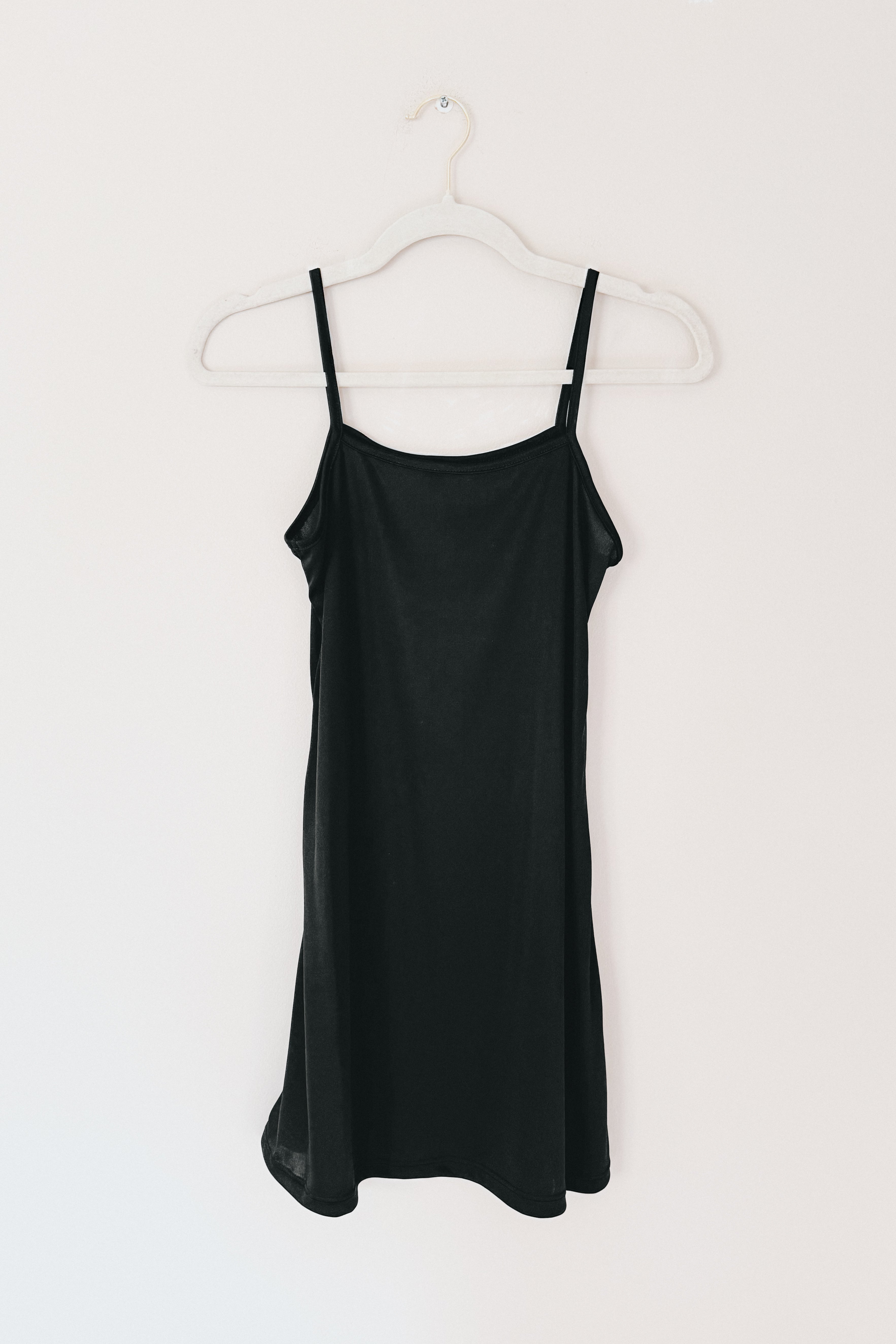 Simple black slip dress with thin adjustable straps hanging on white hanger against plain wall