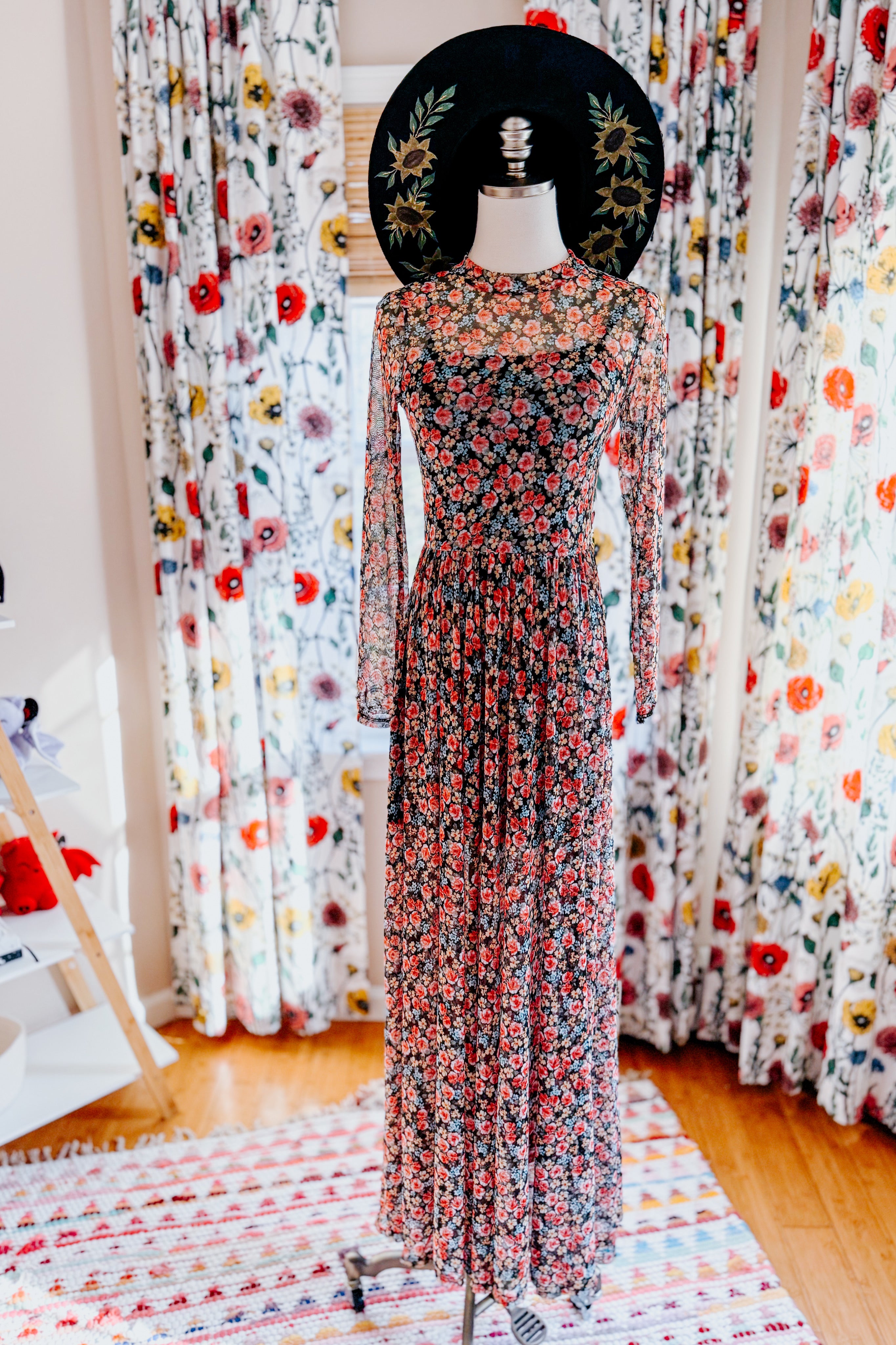 Floor-length floral print mesh dress with long sleeves styled on mannequin in a bright room with floral curtains and rug
