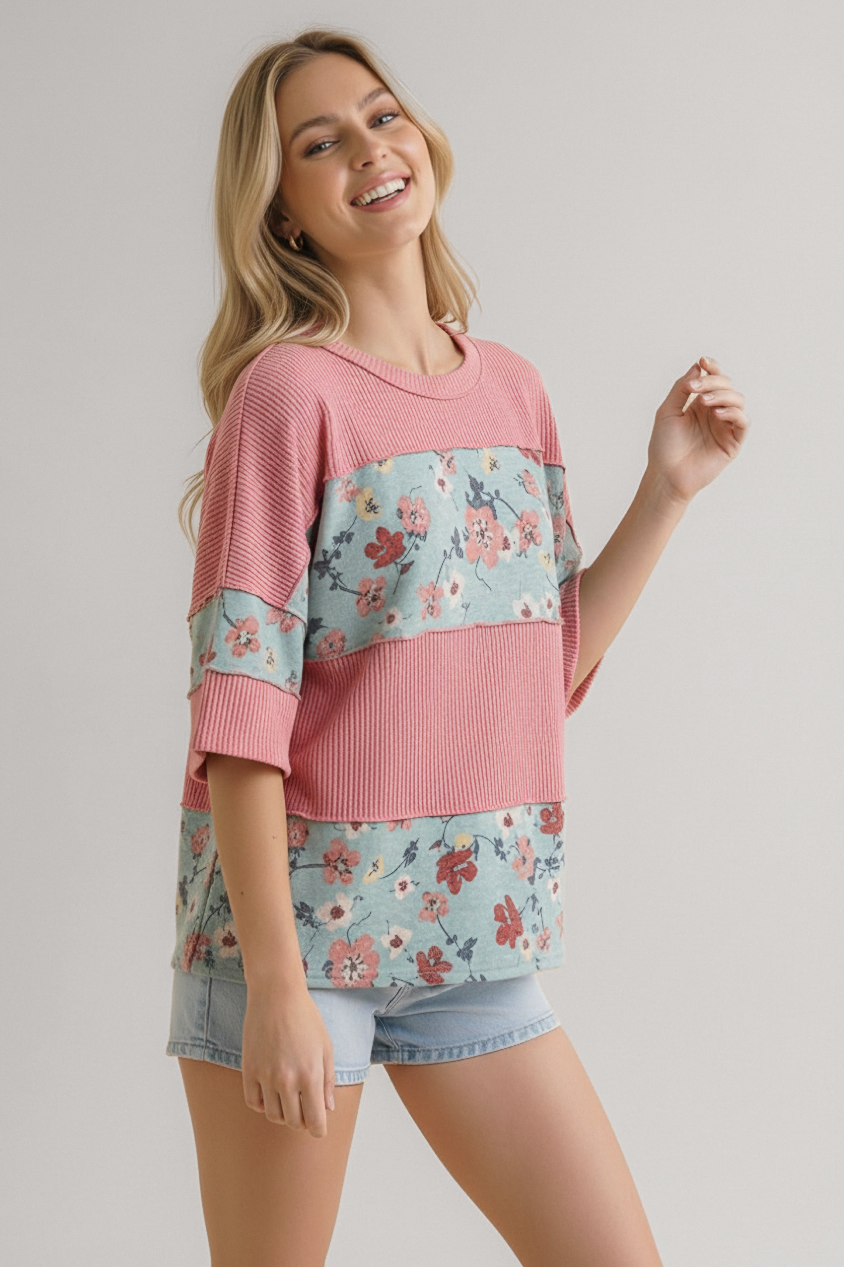 Woman wearing pink garden party floral colorblock top with elbow-length sleeves and light denim shorts smiling against gray background