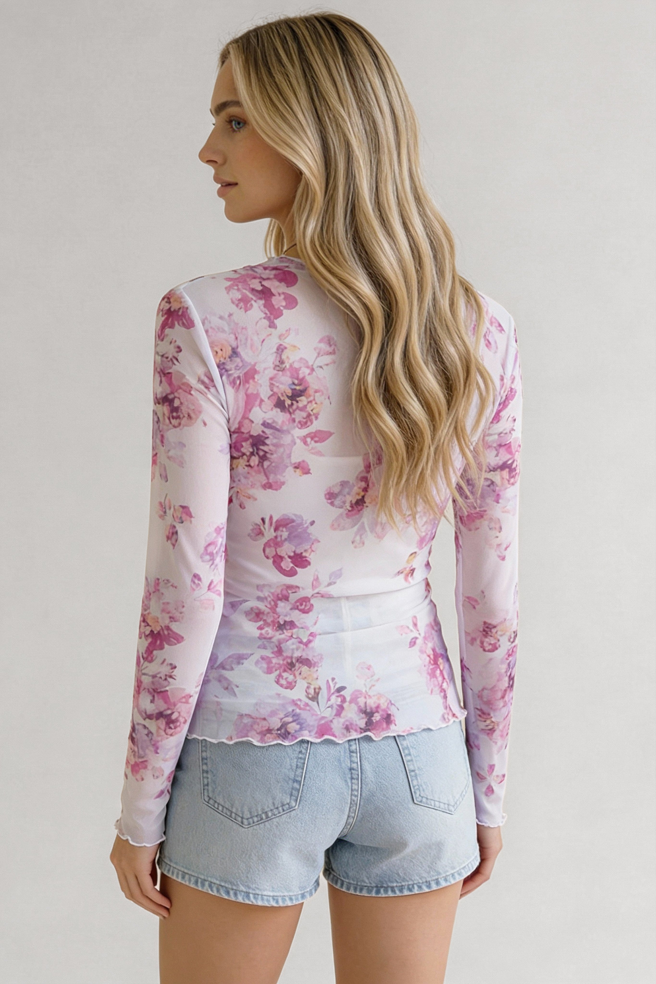Woman wearing pink floral sheer long-sleeve top with lettuce-edge hem paired with light denim shorts shown from back