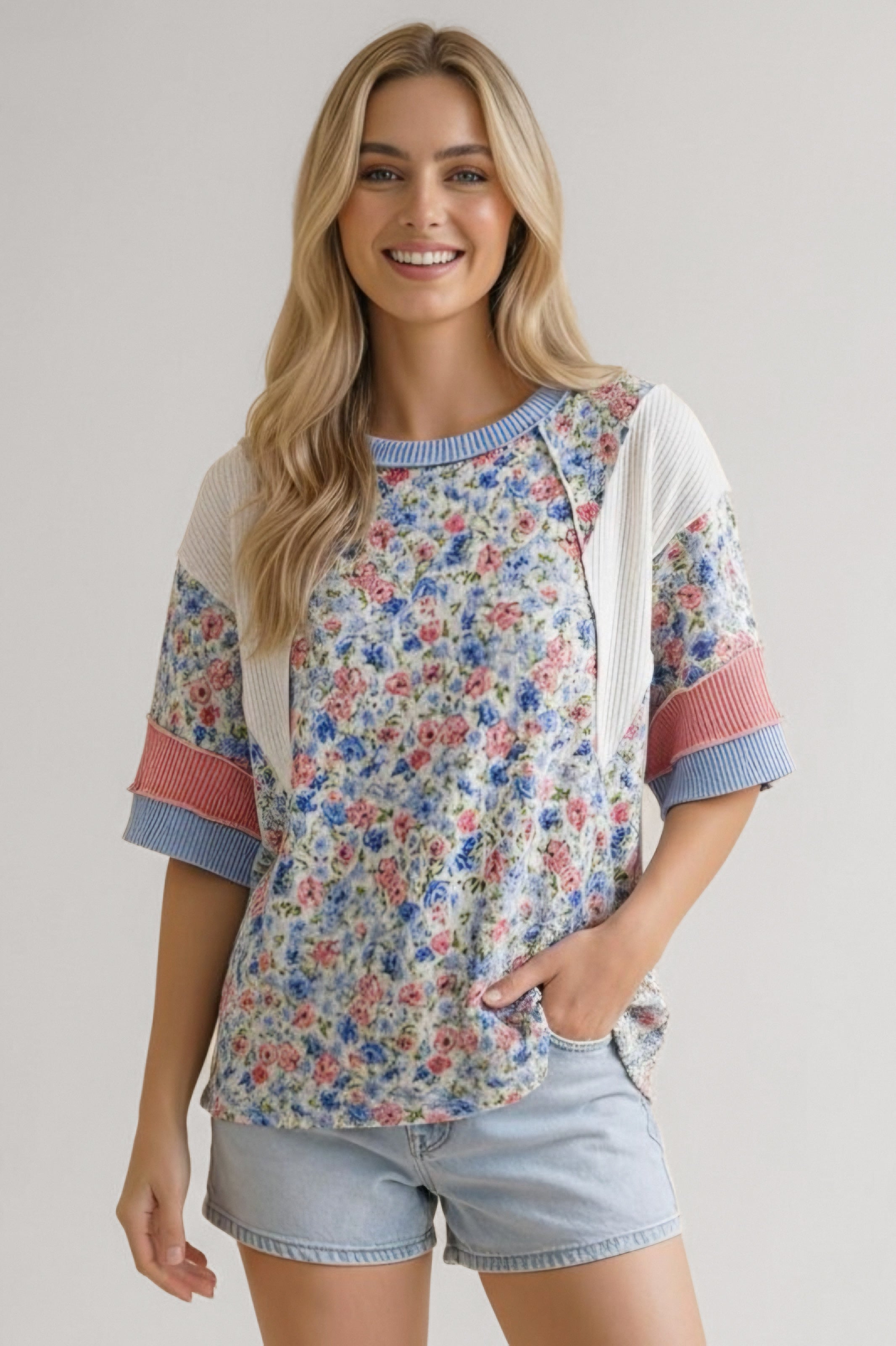 Smiling woman wearing mixed media floral top with blue accents and light denim shorts against neutral background