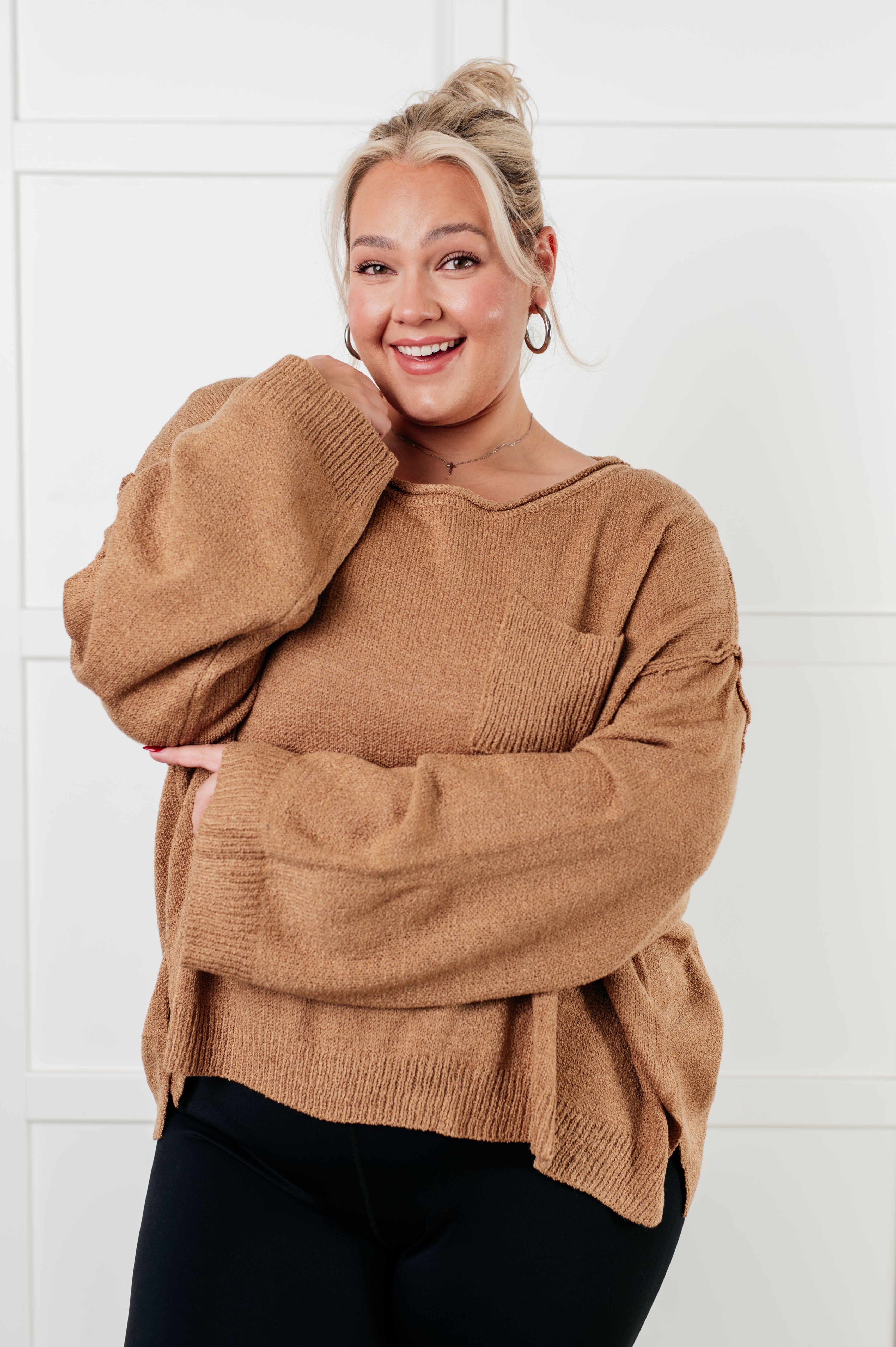Smiling plus size woman in loose camel knit boatneck sweater with ribbed cuffs and front pocket against white background