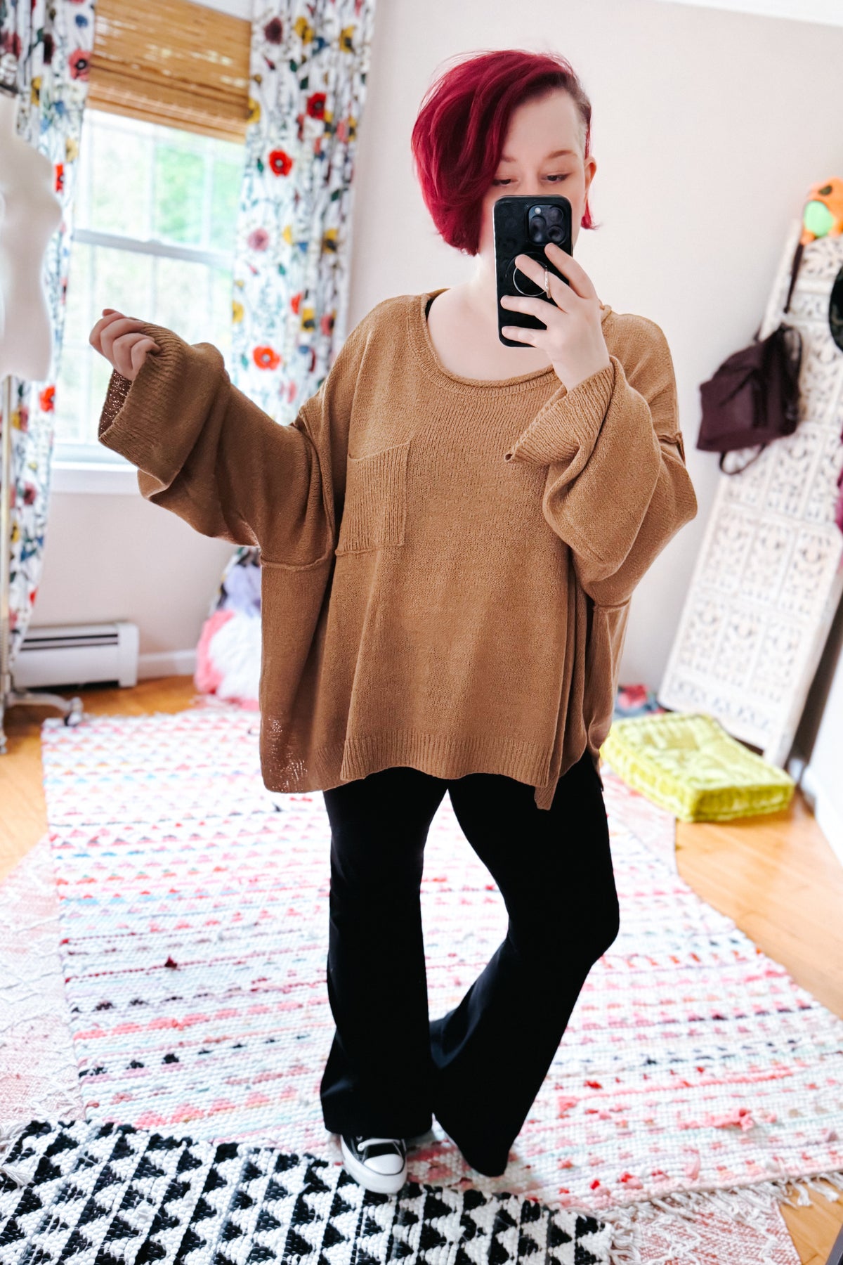 Red haired woman taking a bedroom mirror selfie in a camel boatneck sweater with chest pocket black flared pants and white sneakers