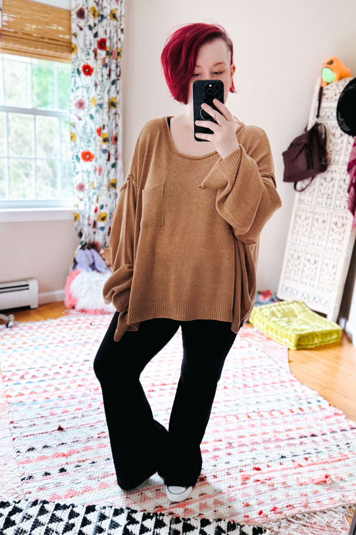 Red haired woman taking a bedroom mirror selfie in a camel boatneck sweater with chest pocket, black pants, and white sneakers