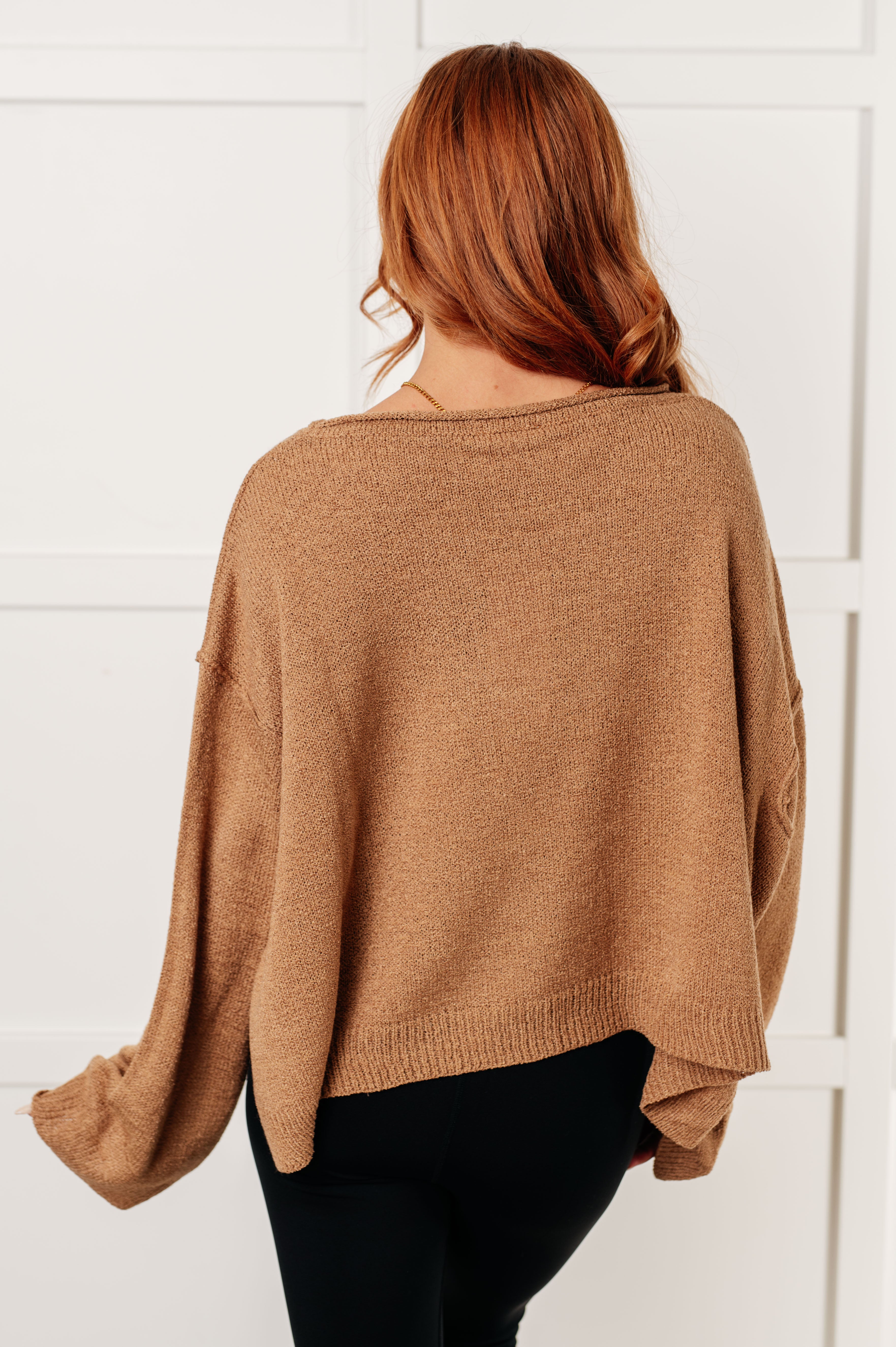 Woman with wavy auburn hair wearing loose camel boatneck sweater with long sleeves shown from the back