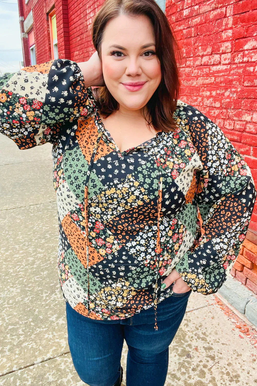 Woman wearing a floral patchwork top with long sleeves and a front tie standing against a red brick wall outdoors