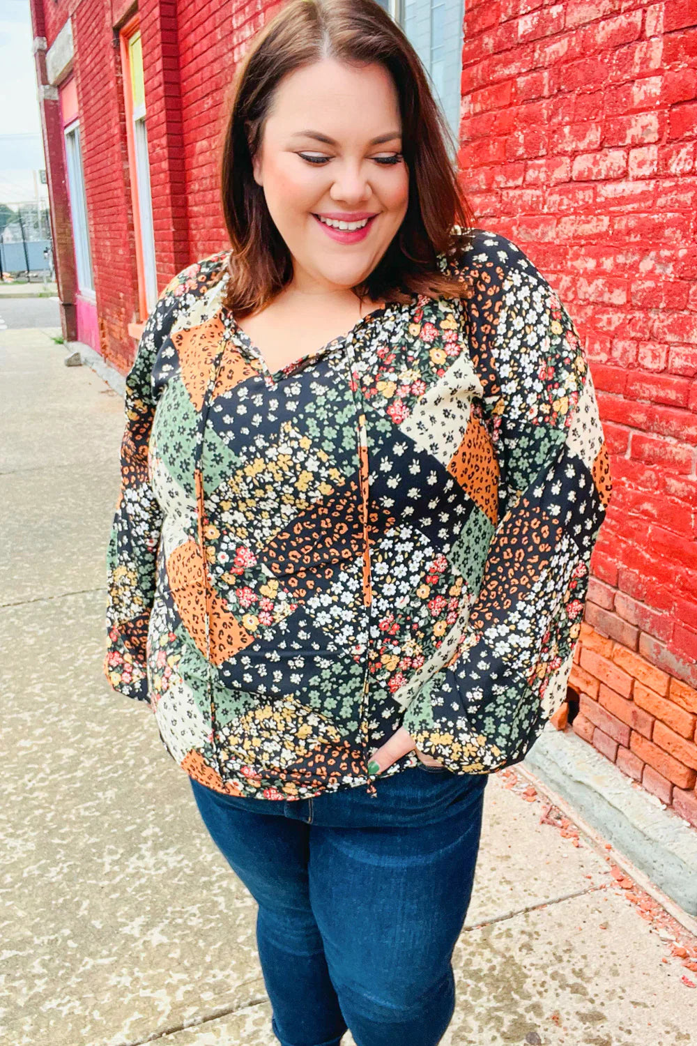 Smiling woman outdoors wearing a green and brown floral patchwork top with a keyhole tie neckline and dark jeans