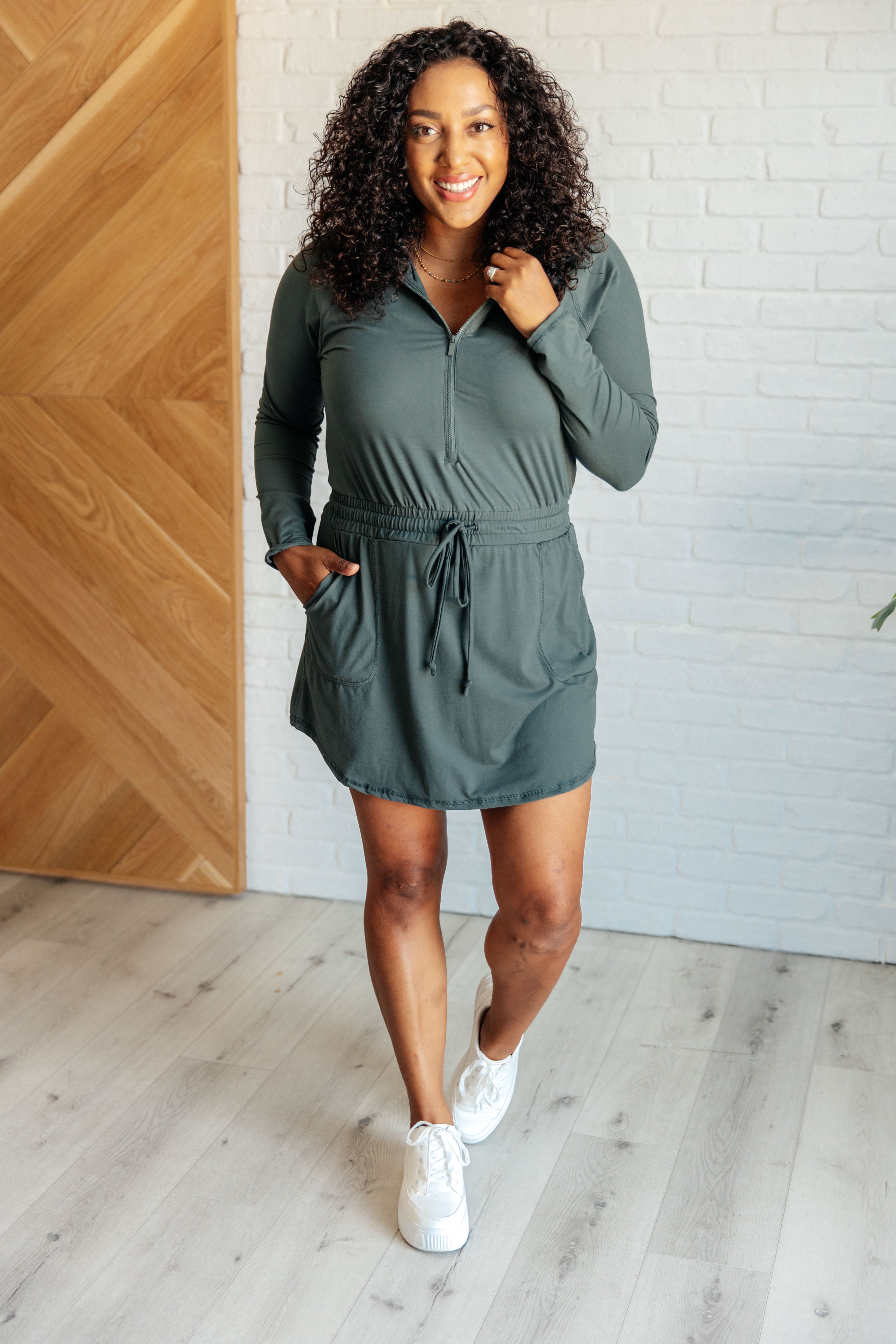 Woman with curly hair wearing a smoked spruce long sleeve hoodie romper with front pockets and drawstring waist standing indoors smiling