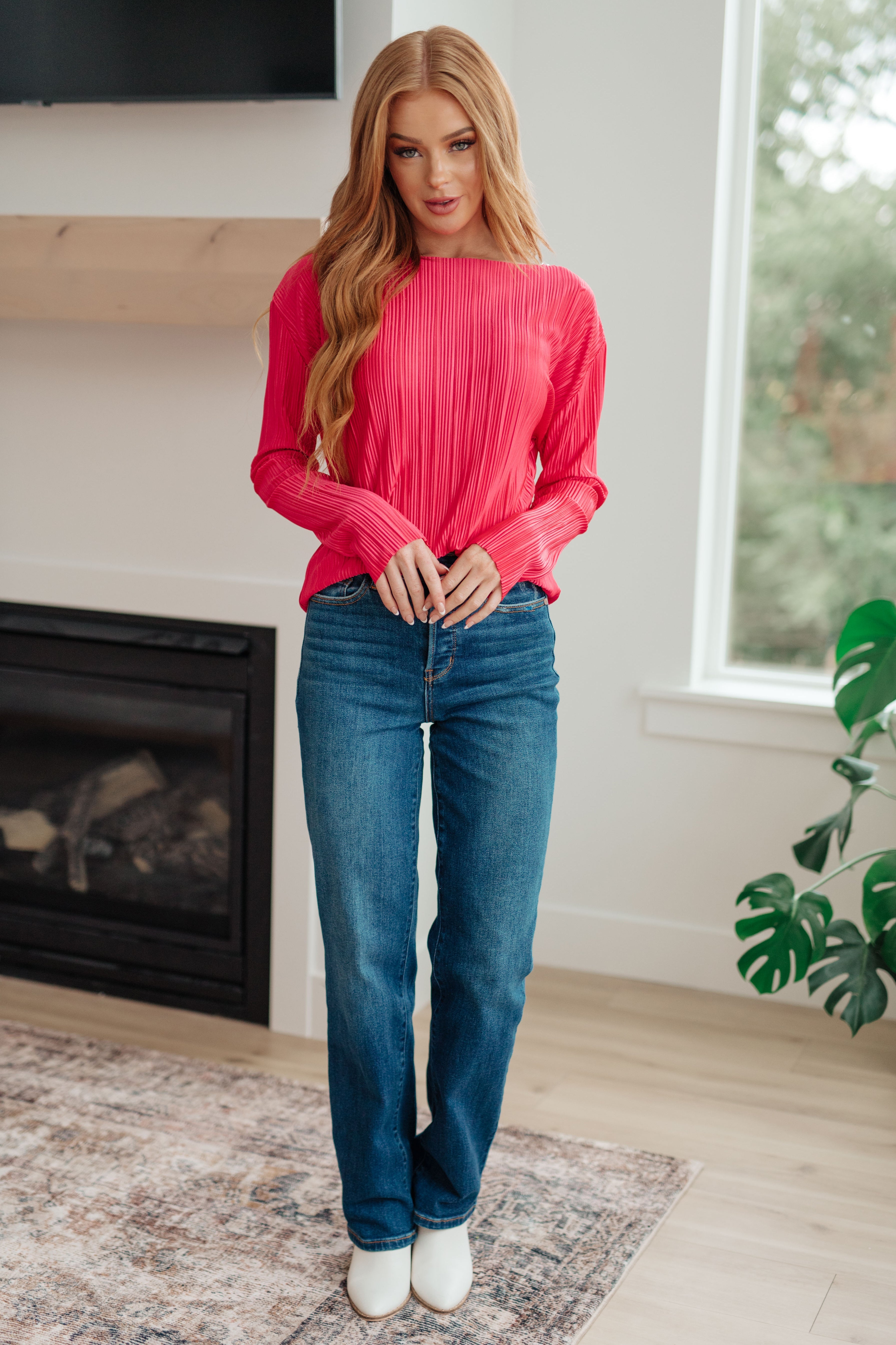 Woman wearing a bright pink blouse, blue jeans and white boots in an indoor setting 