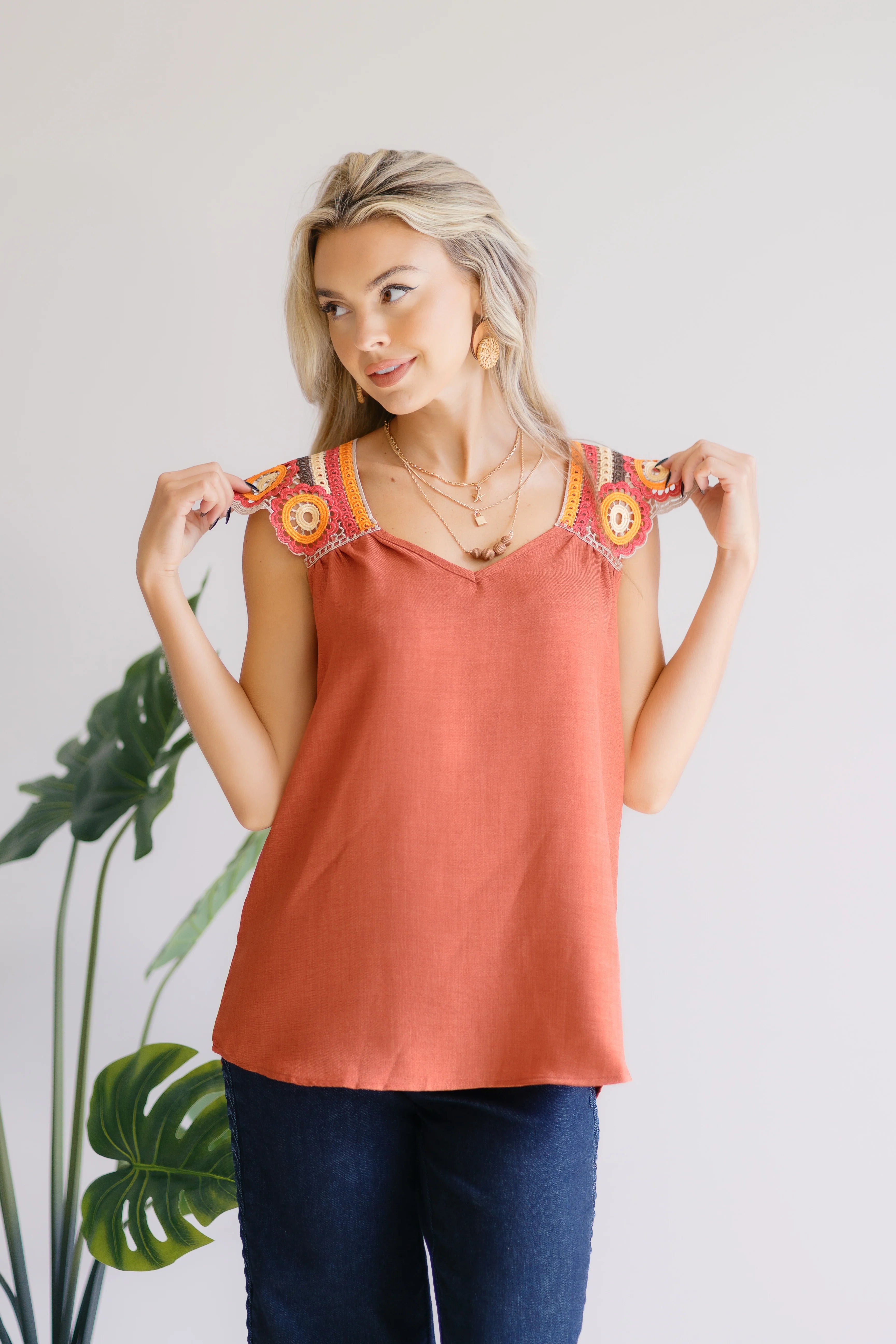 Woman wearing a rust orange sleeveless top with colorful crochet shoulder panels and dark blue jeans posing indoors