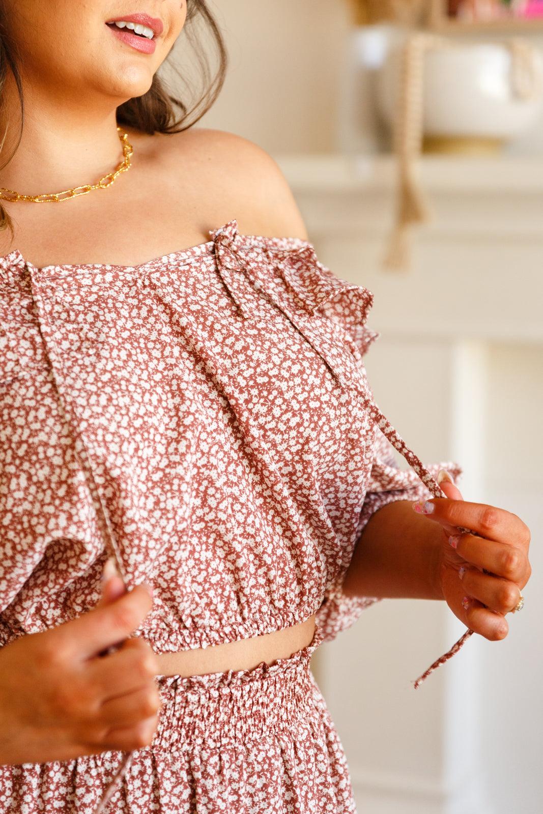 Close-up of woman adjusting rose-colored floral off-shoulder crop top with ruffle and tie details indoors