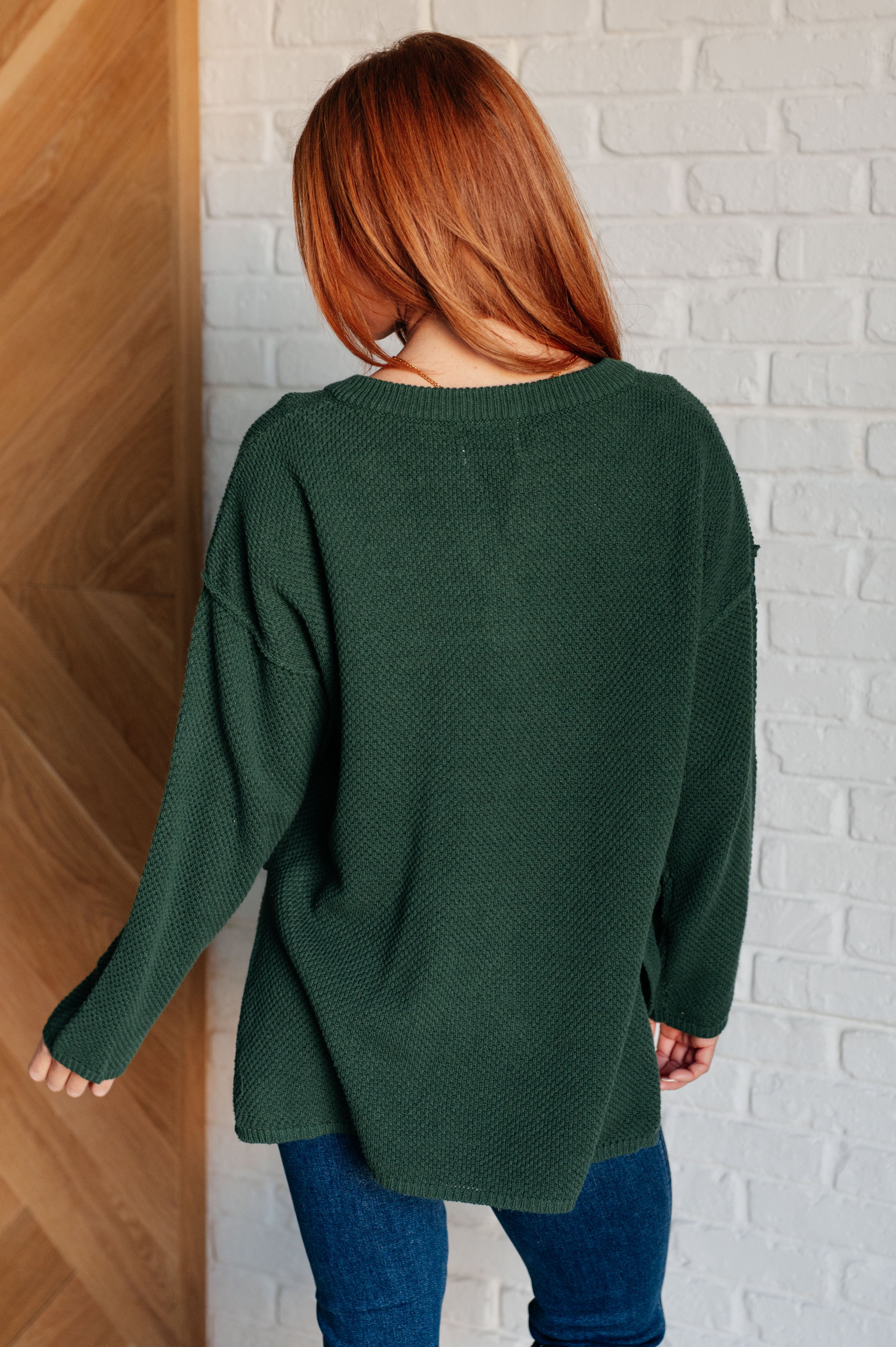 Back view of woman wearing deep green knit henley sweater with relaxed fit paired with dark blue jeans indoors
