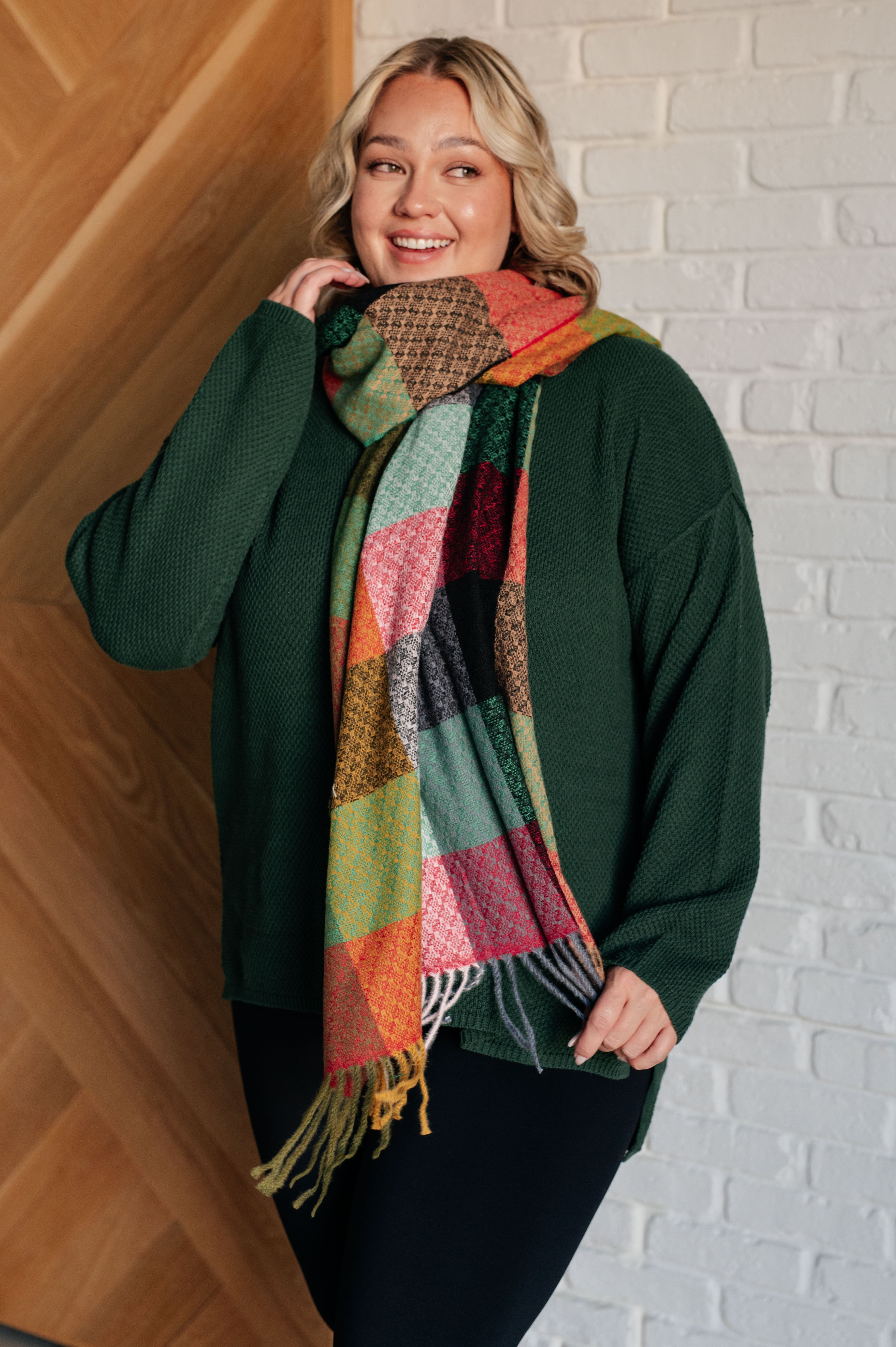 Smiling woman wearing dark green textured henley sweater paired with colorful plaid scarf against white brick wall