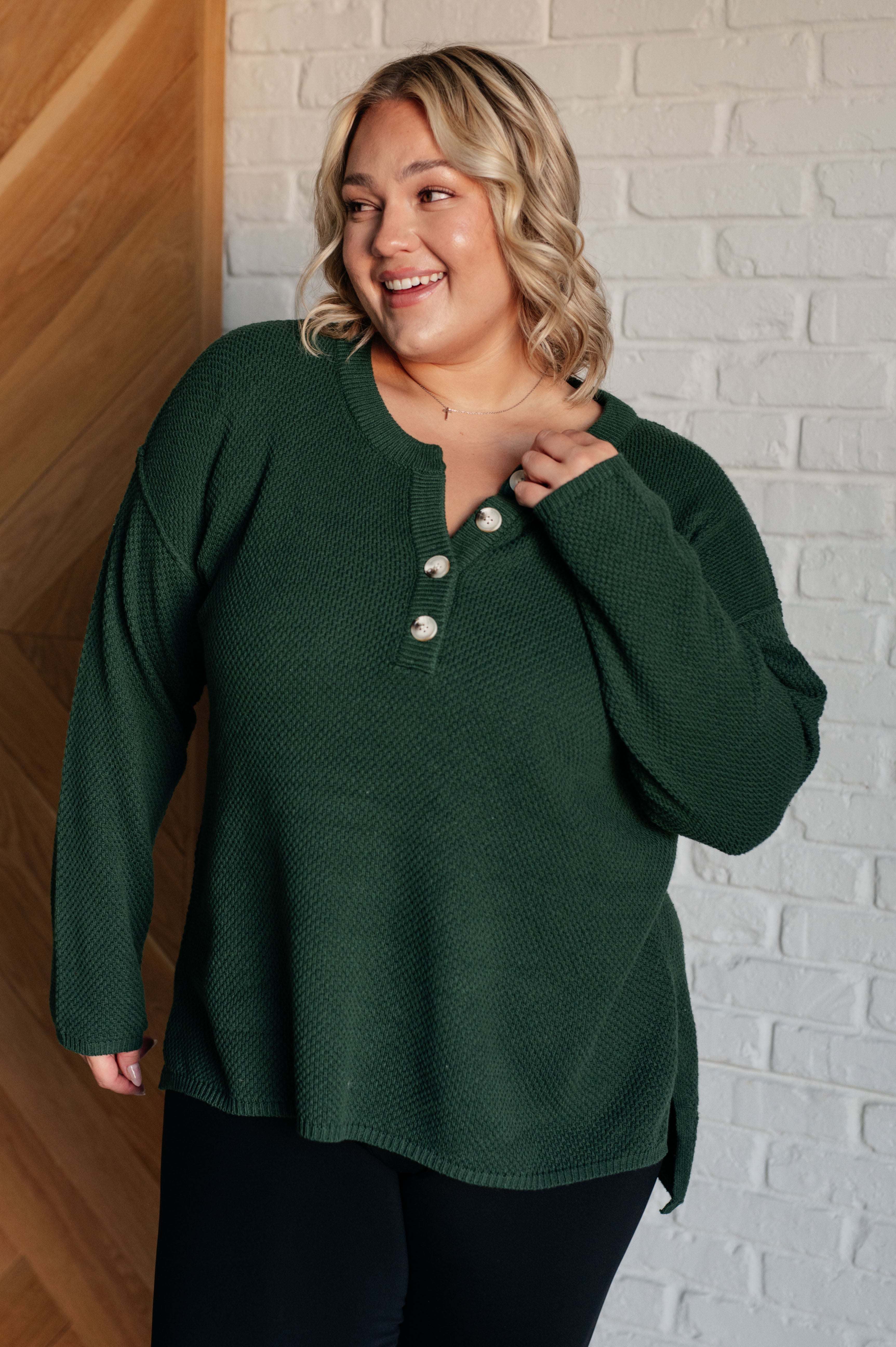 Smiling woman wearing dark green waffle knit henley sweater with button placket and black pants indoors
