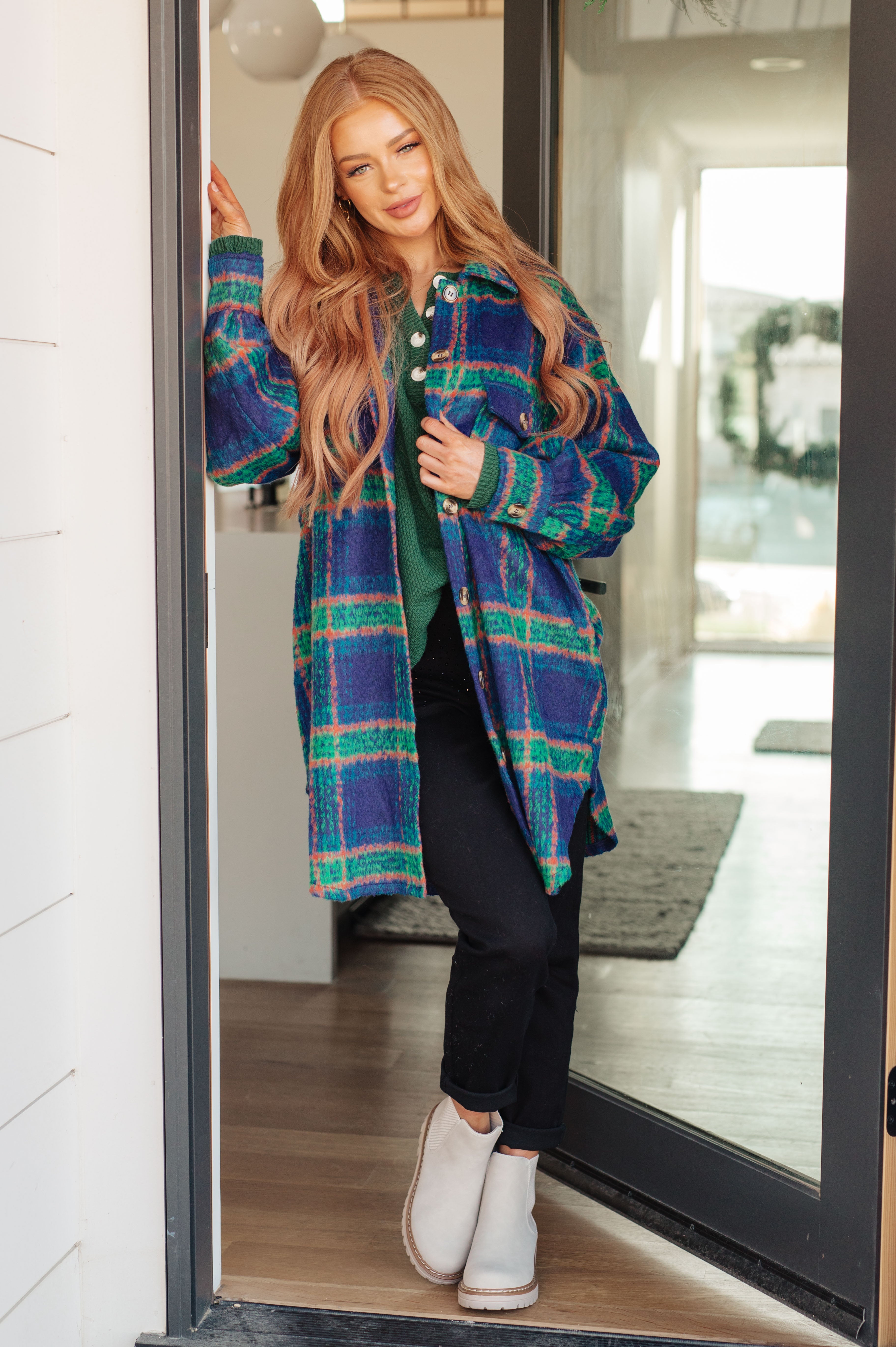 Woman standing in doorway wearing green henley sweater under blue and green plaid long jacket with black pants and white boots