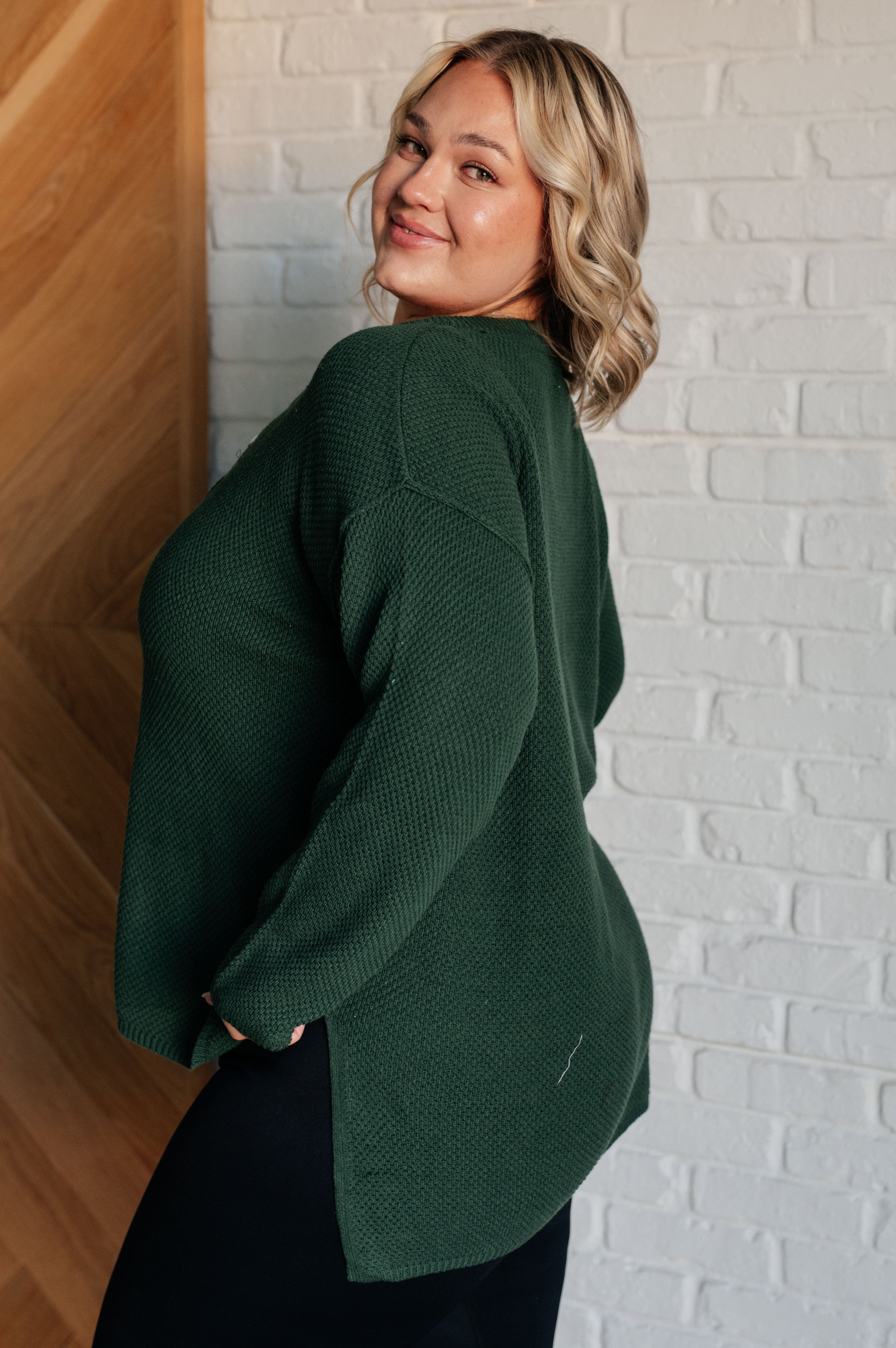Smiling woman wearing loose dark green textured knit henley sweater standing against white brick wall
