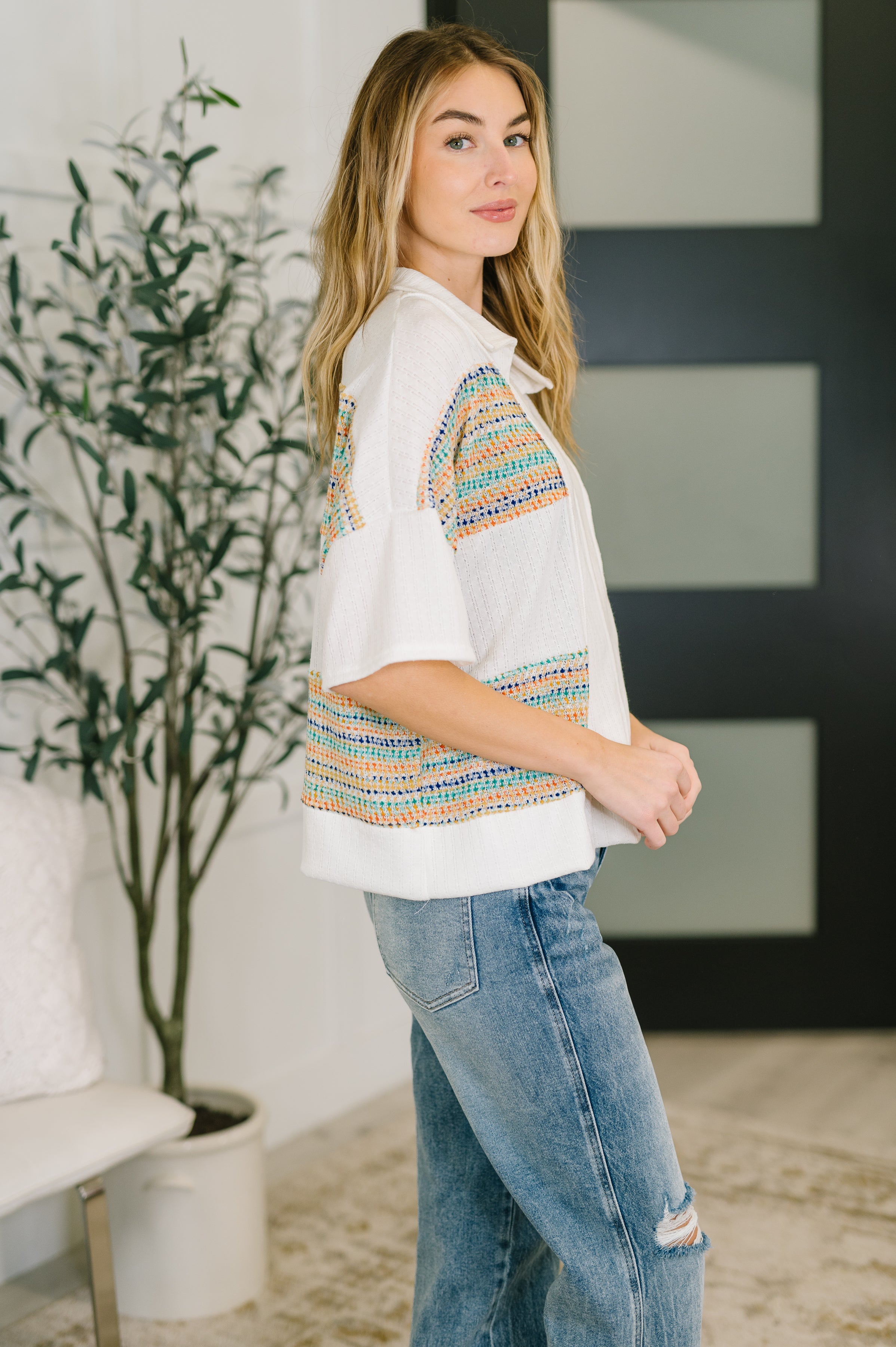 Woman wearing an ivory knit top with colorful woven panels and flared sleeves paired with distressed blue jeans indoors