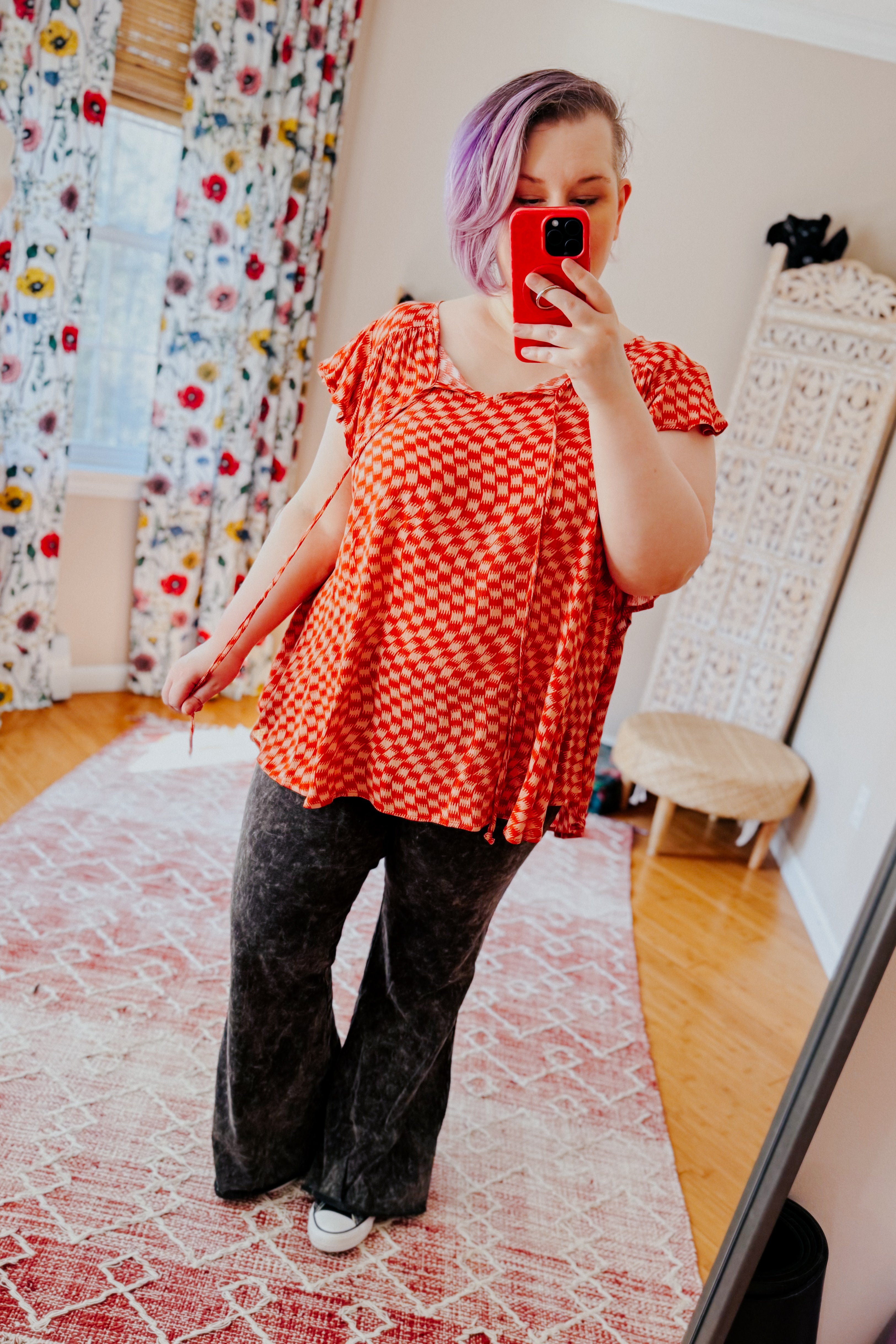 Woman with short purple hair wearing orange houndstooth butterfly sleeve top and dark wide-leg pants taking mirror selfie indoors