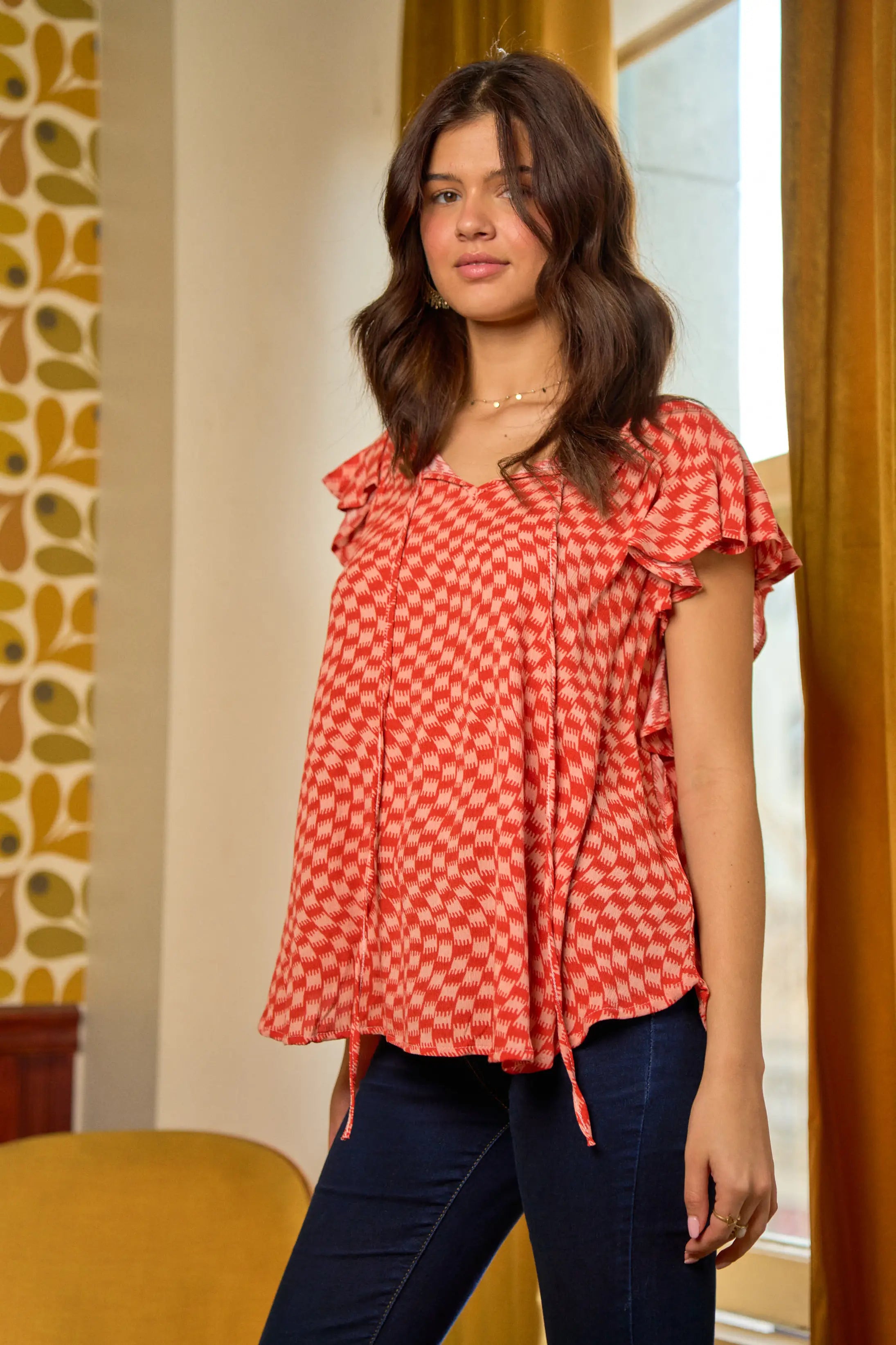 Young woman wearing red-orange houndstooth top with flutter sleeves standing indoors by window with mustard curtains