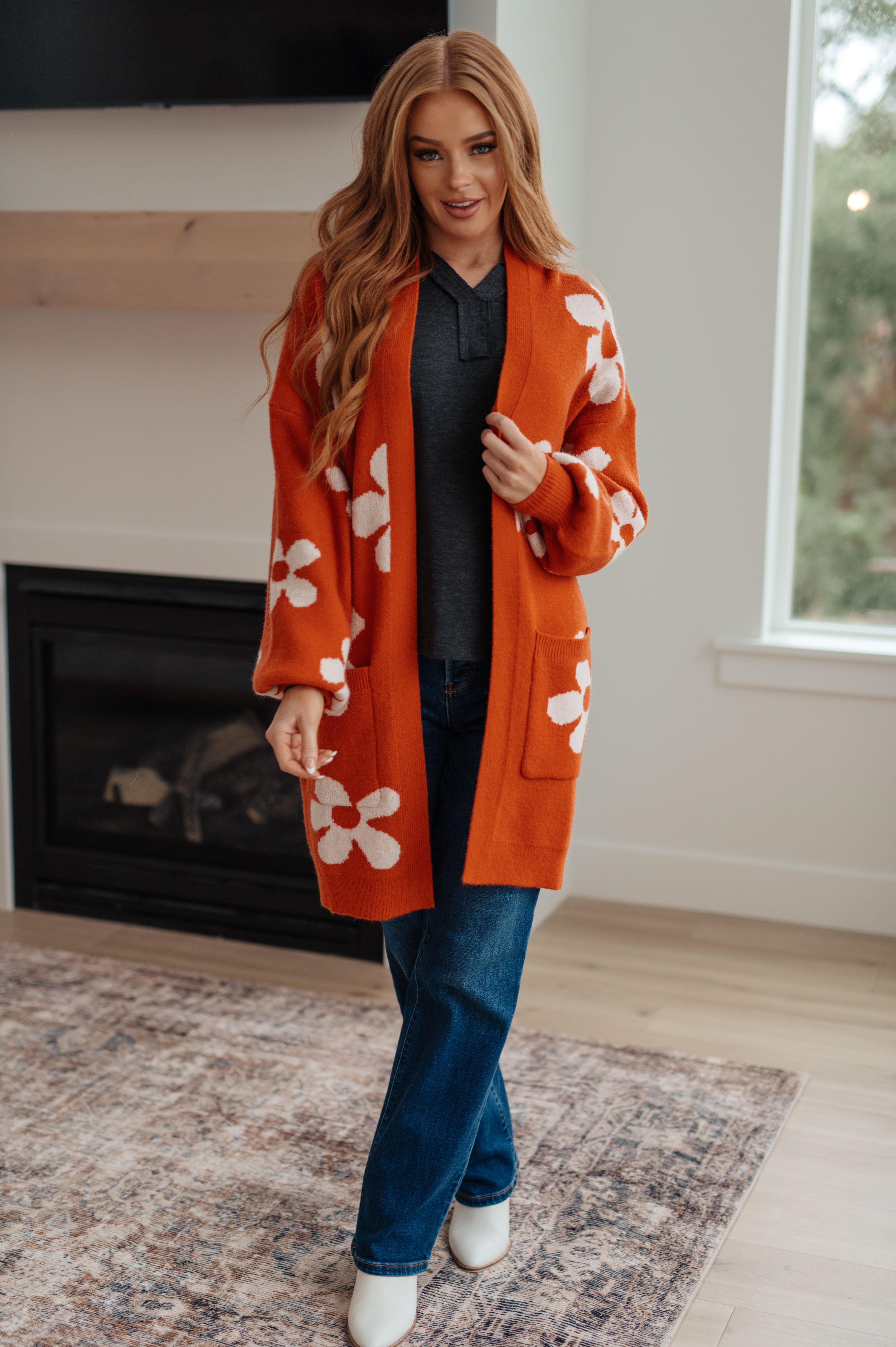 Woman wearing charcoal long sleeve Good Luck Charm top under orange floral cardigan and blue jeans in modern living room