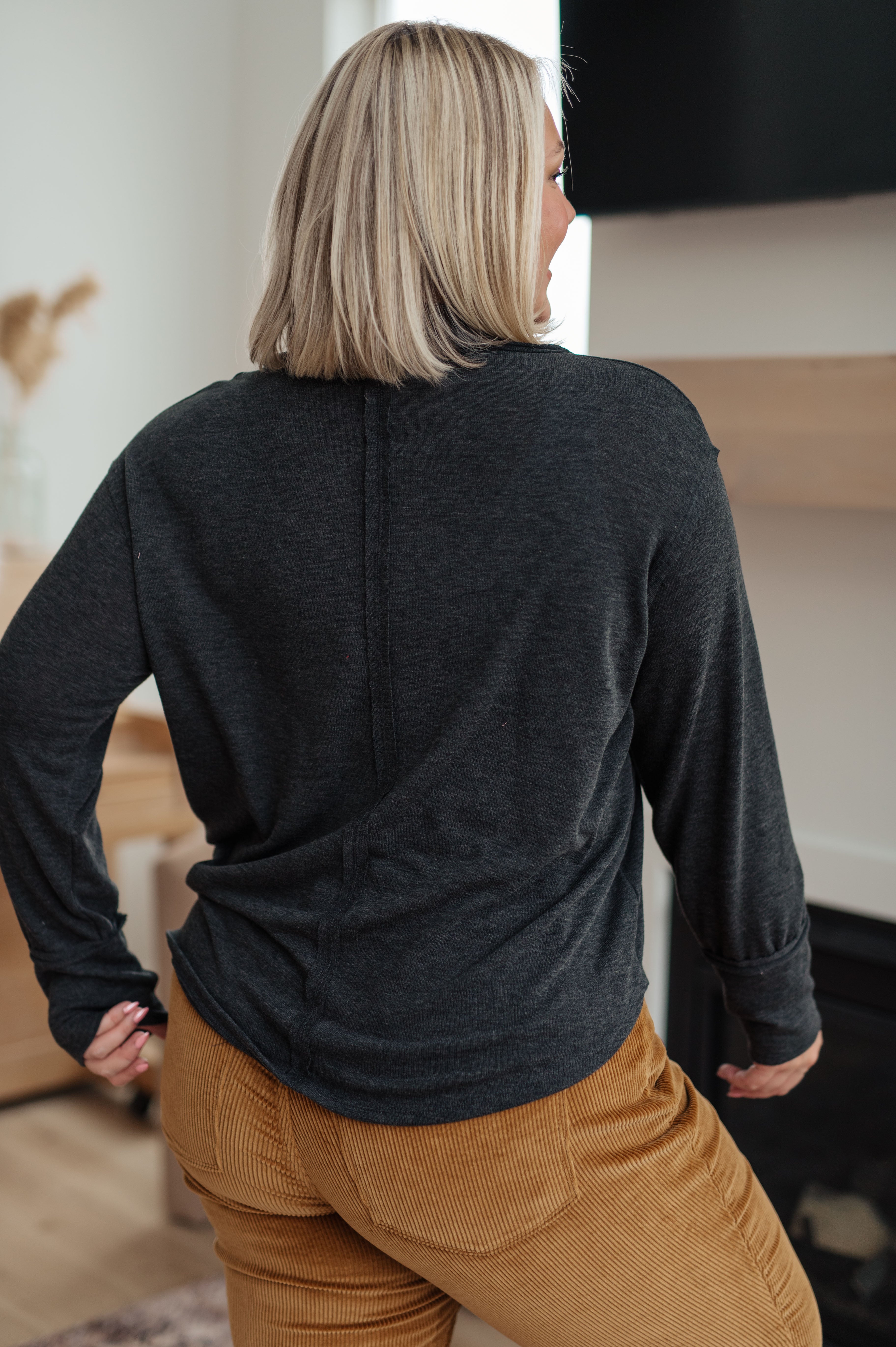Woman seen from behind wearing charcoal long sleeve top with seam detail paired with brown corduroy pants in cozy room