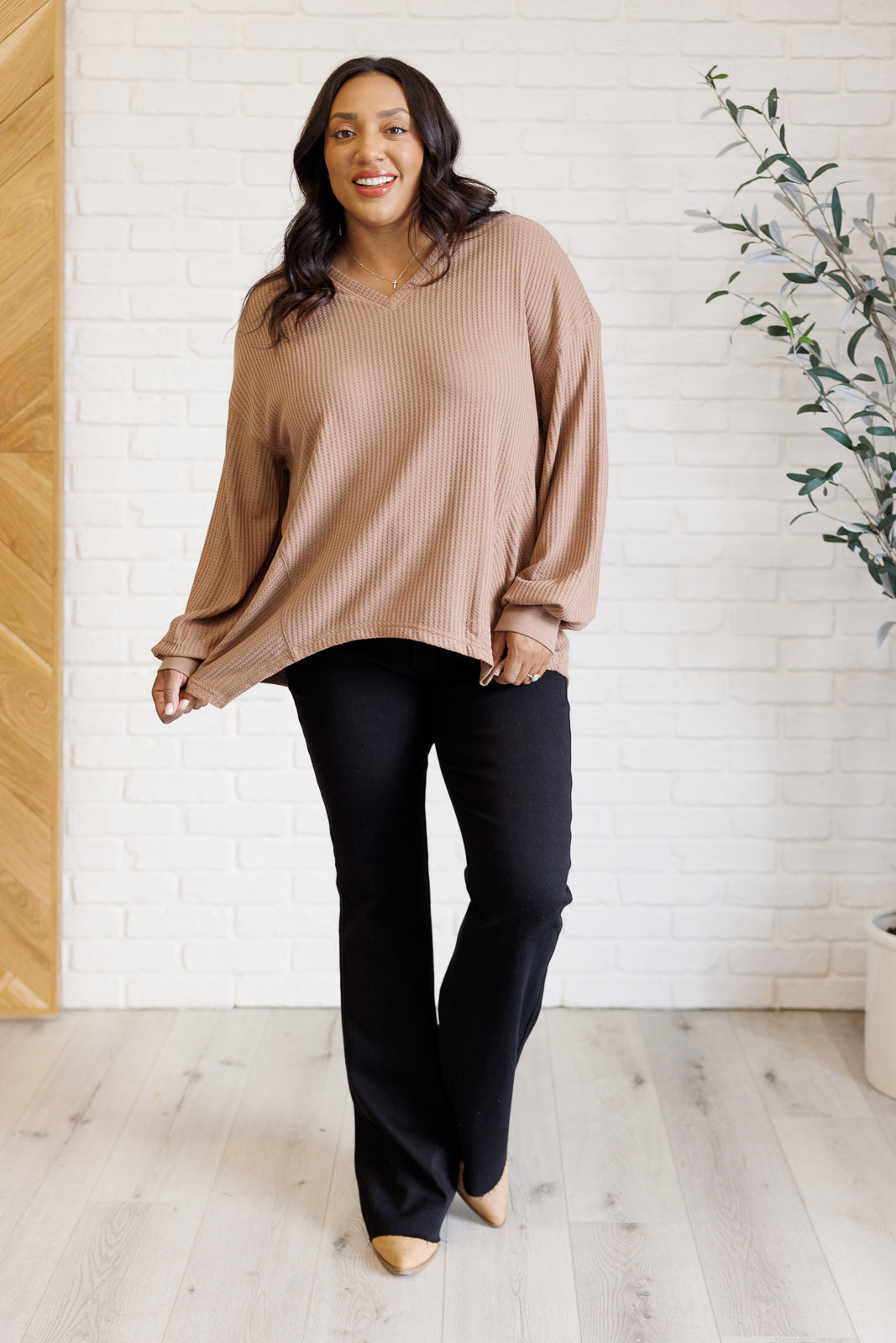 Woman wearing a light brown waffle knit v neck long sleeve top, black boot leg jeans and tan boots standing in front of a white brick wall.