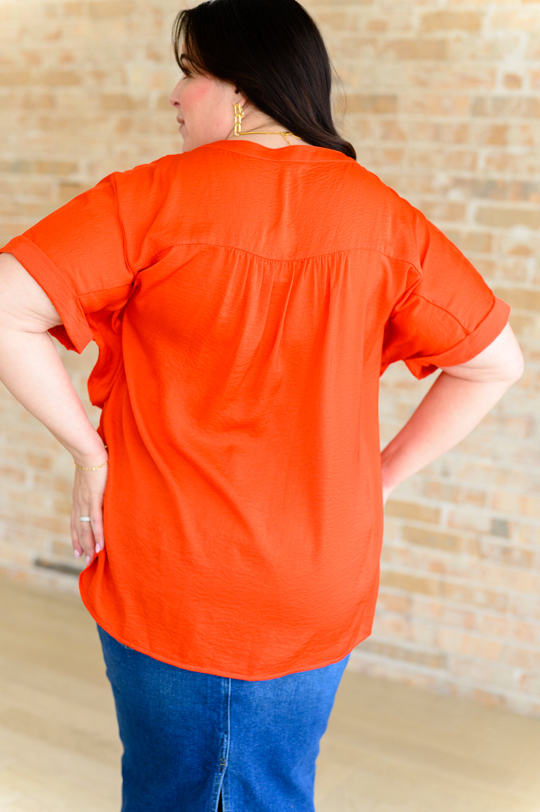 Plus size woman modeling vibrant orange blouse with short sleeves and gathered back detail against brick wall
