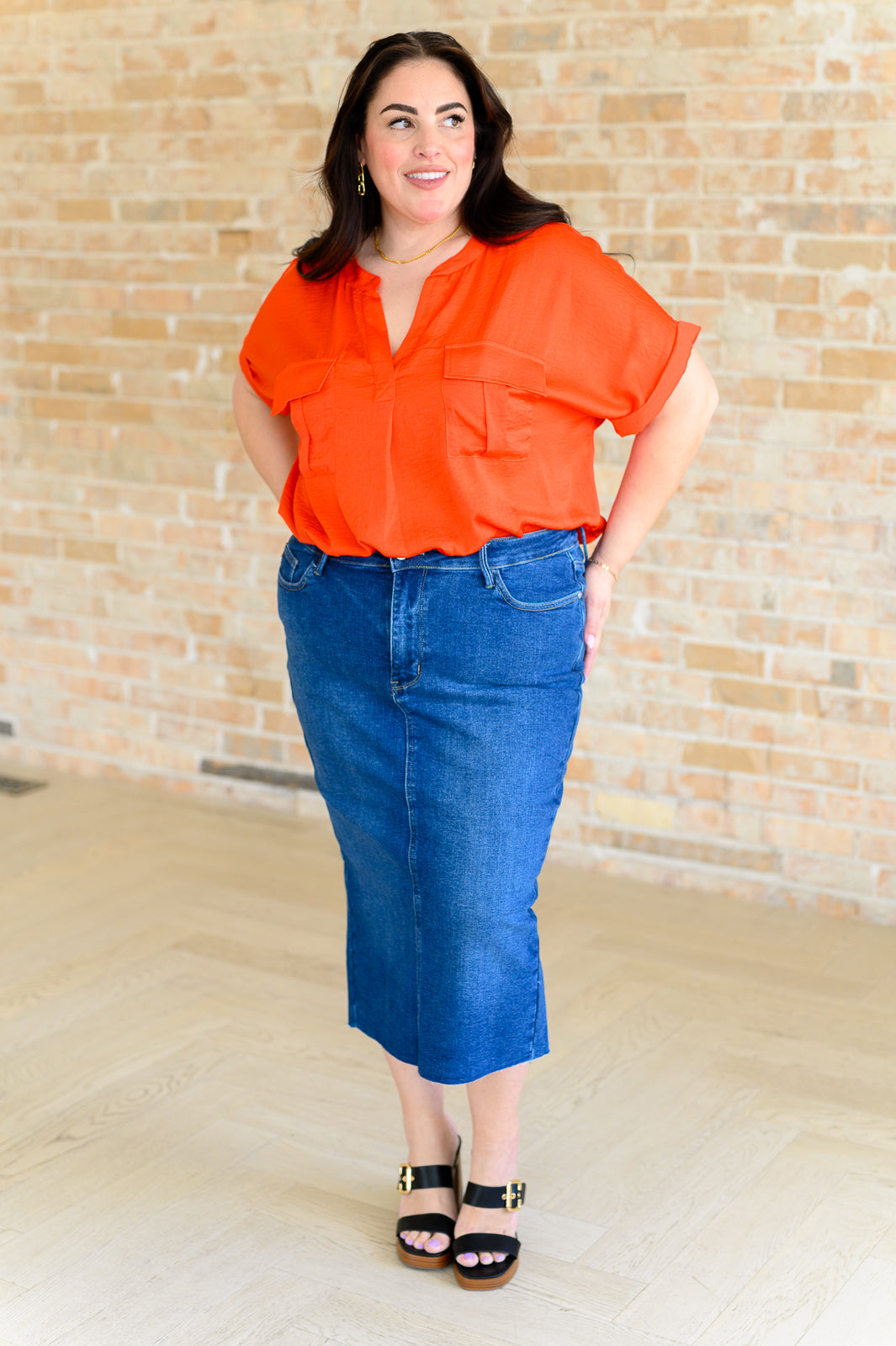 Woman wearing vibrant orange blouse with rolled sleeves paired with knee-length denim skirt and black sandals