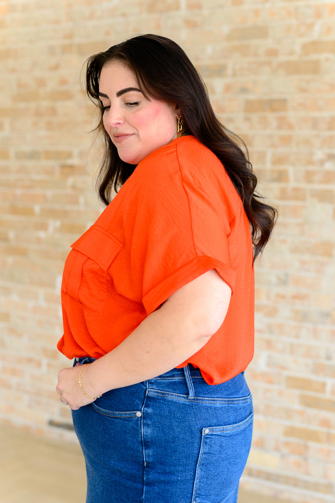 Woman wearing vibrant orange blouse with rolled sleeves paired with blue denim jeans against a brick wall background