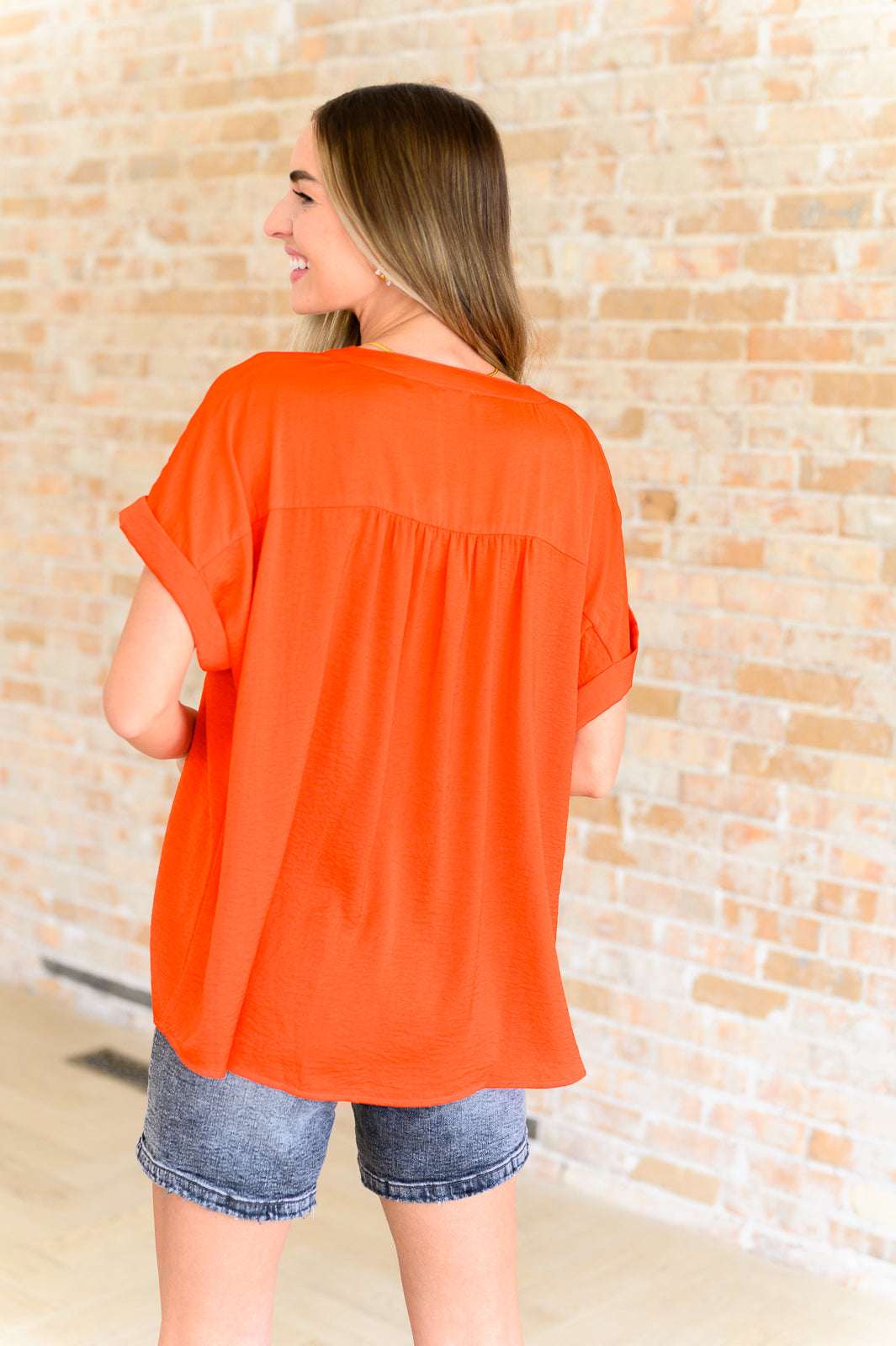 Woman shown from behind wearing loose bright orange blouse with rolled sleeves and faded denim shorts indoors