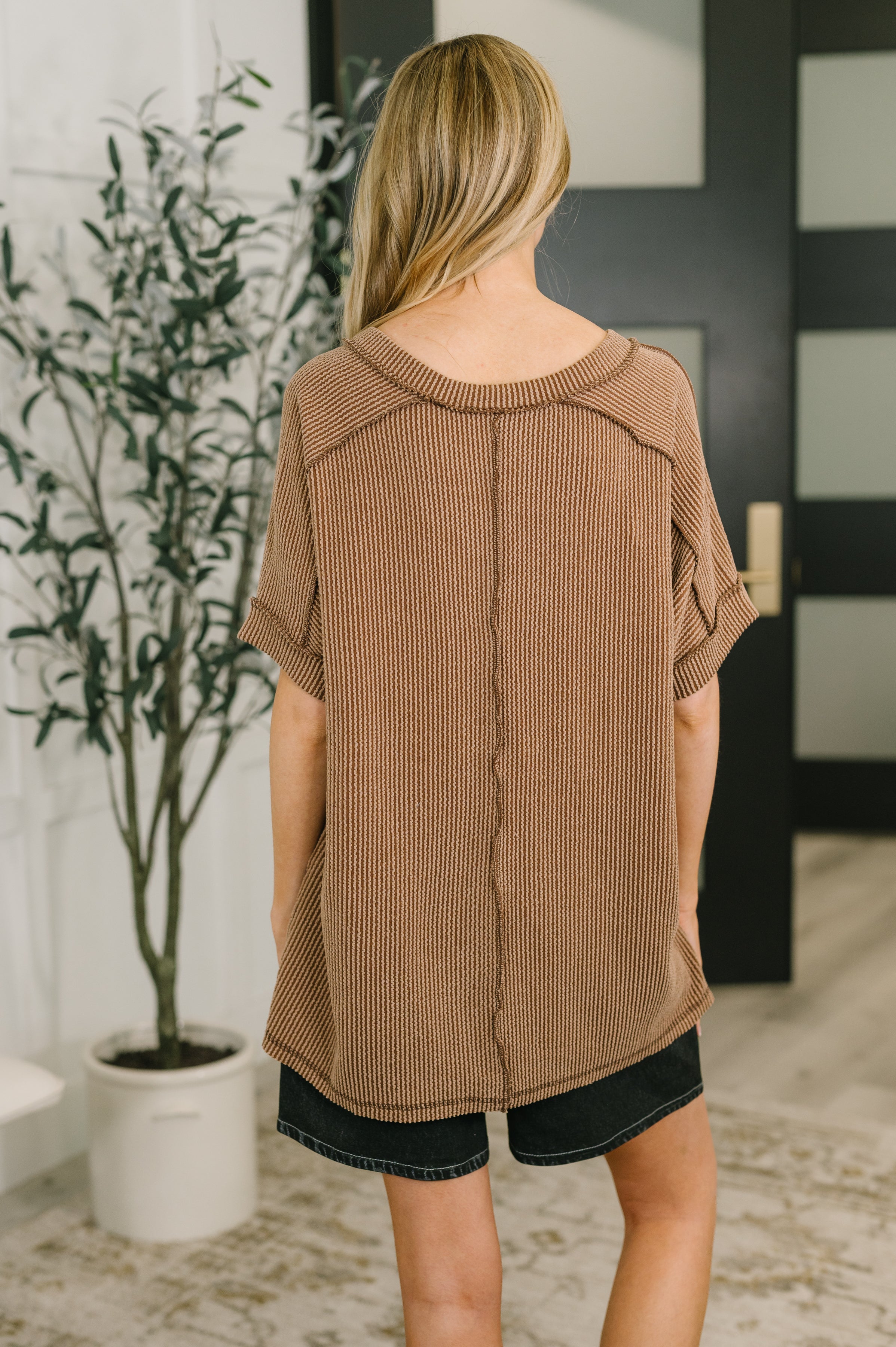 Back view of a woman wearing a brown textured knit top with rolled sleeves paired with black denim shorts indoors