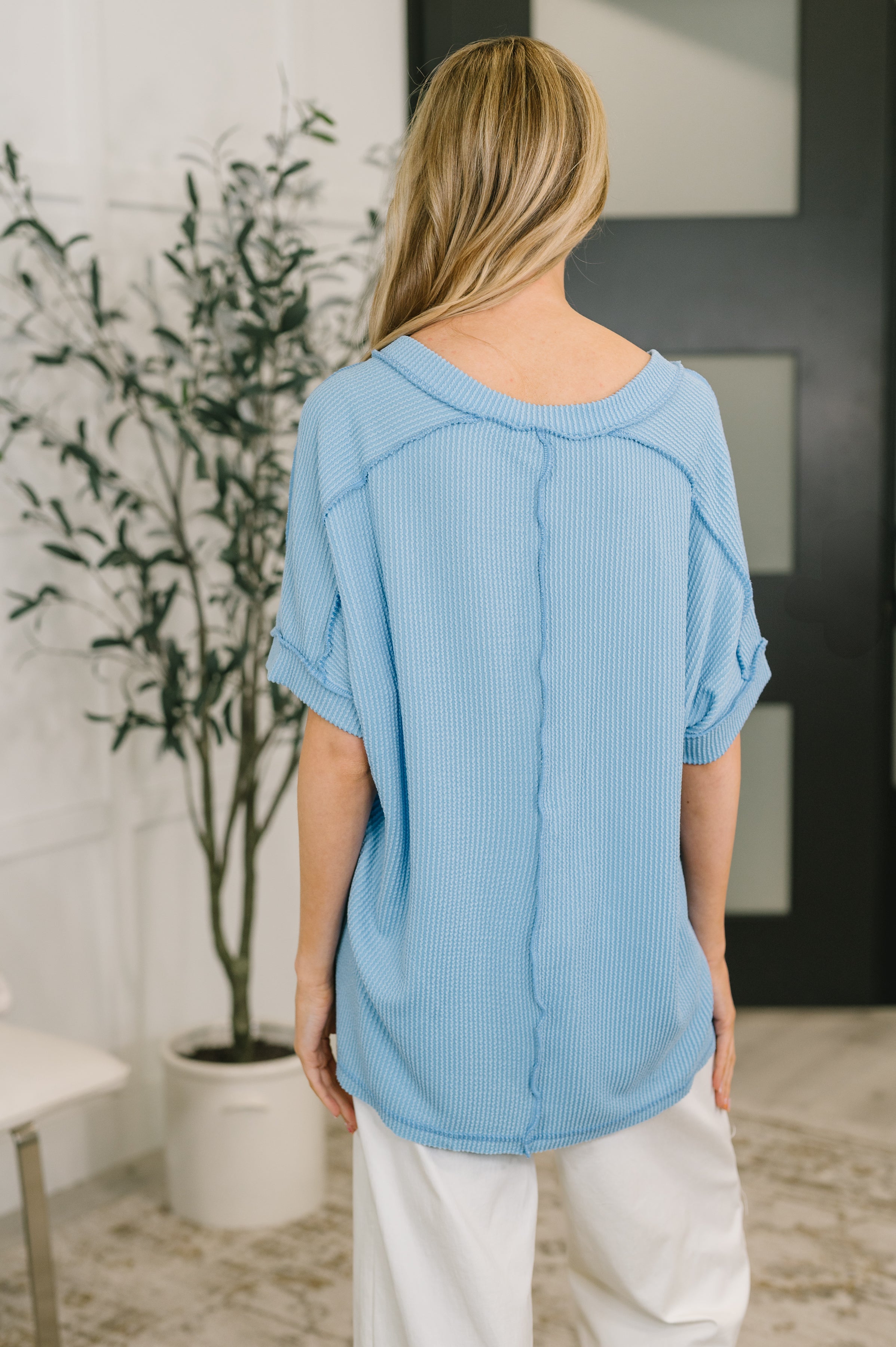 Woman seen from behind wearing a spring blue textured knit top with rolled short sleeves and white pants indoors
