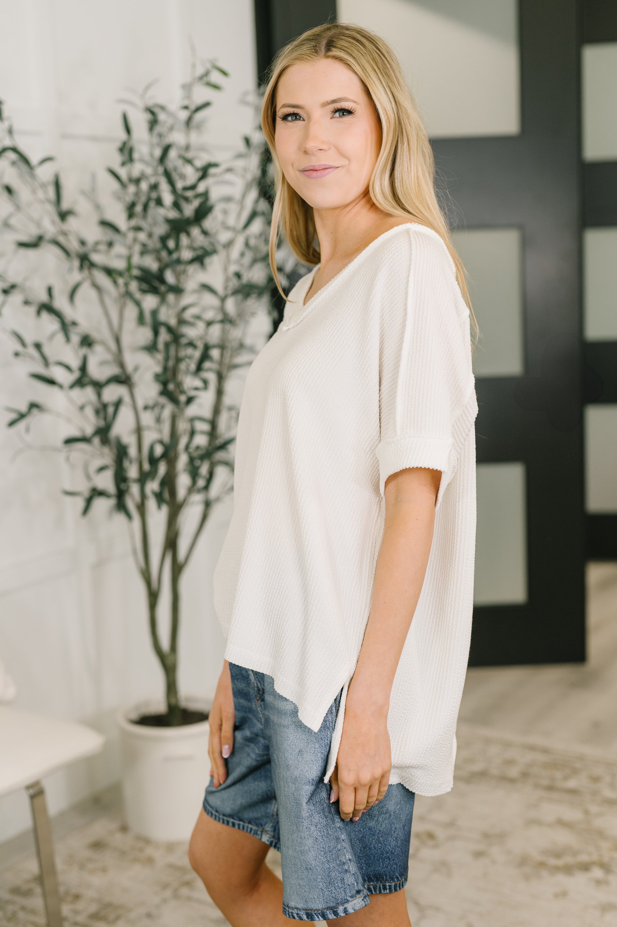 Woman smiling indoors wearing a loose white textured V-neck top with short cuffed sleeves and denim shorts