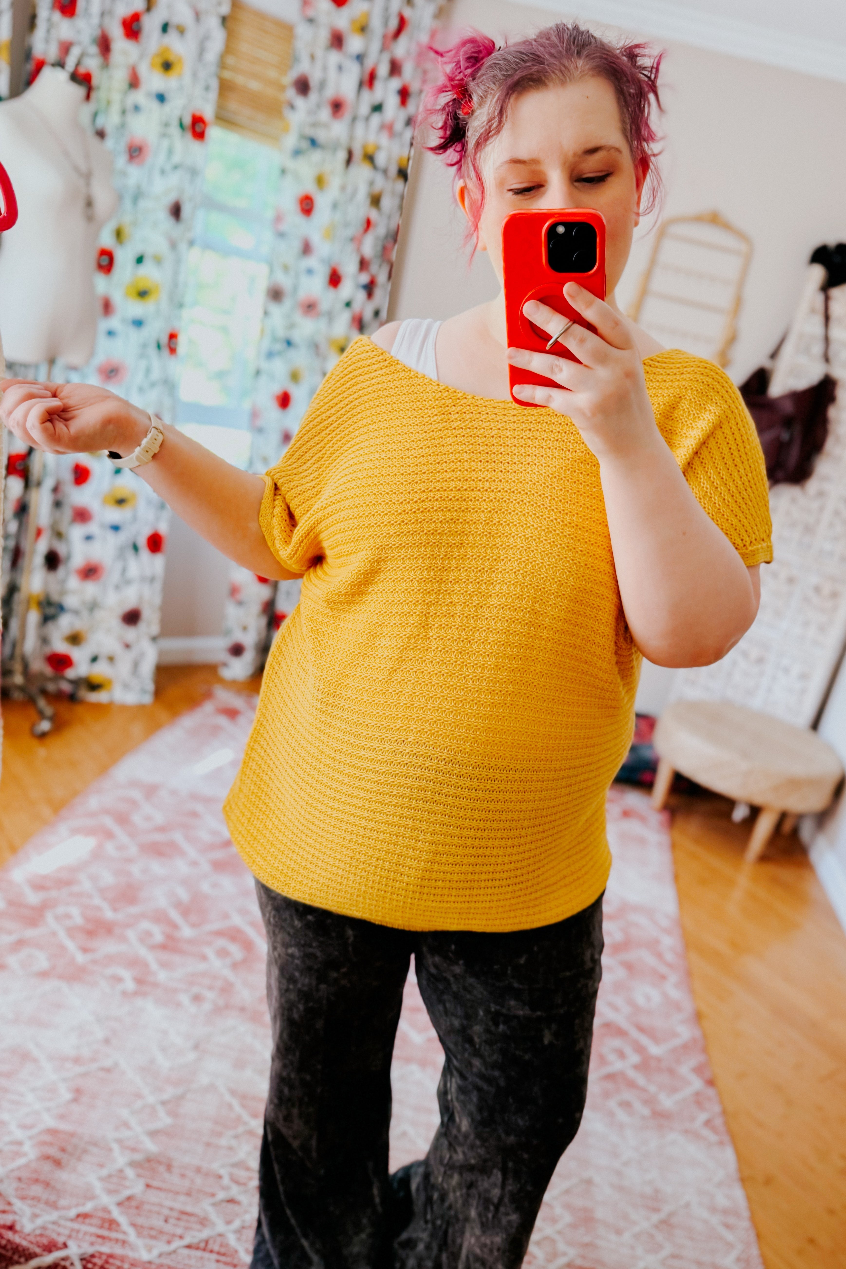 Woman taking selfie wearing loose textured yellow knit top with wide neckline and black pants in cozy room with floral curtains